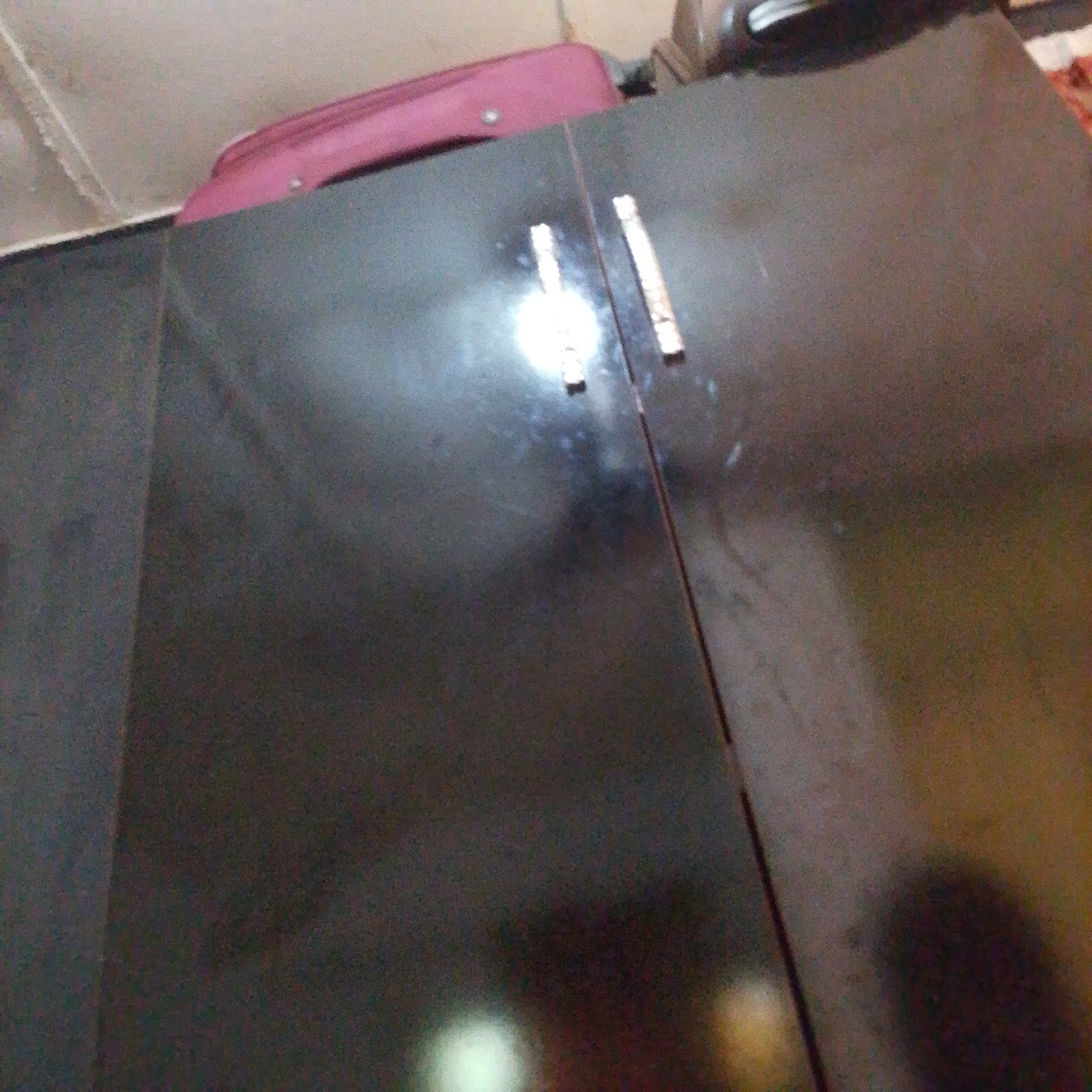 The image shows a close-up, angled view of a dark, glossy surface, likely the top of a piece of furniture such as a cabinet or desk. Two small, rectangular metallic objects with etched patterns are visible on this surface, one to the left and one to the right of a central vertical line. These objects appear to be decorative accents or parts of a larger item.
Above the dark surface, a portion of a pink object is visible, which resembles the lid of a suitcase. This object is positioned against a background that appears to be a wall with some texture and a light fixture or architectural detail. The lighting in the image is a mix of ambient and a strong, diffused light source that creates a bright, blurry reflection on the glossy surface.
The overall impression is an indoor setting, possibly a room. The limited visibility of the surroundings and the focus on the dark surface and the pink object suggest a candid or informal capture. No people are visible. The time of day and weather are not discernible from the image. There is no visible text. The location context of Jalingo, Nigeria, does not provide specific visual cues within the image itself.