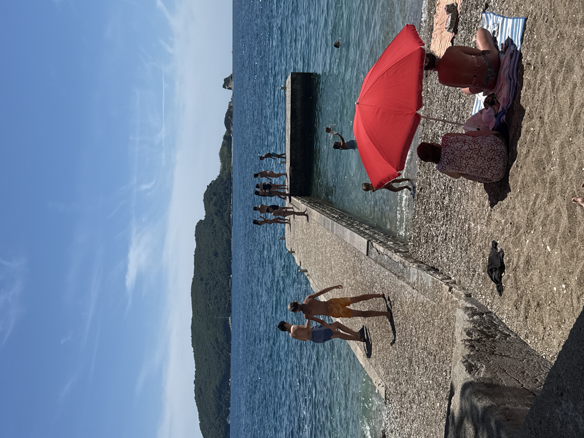 A sunny day at the beach in Budva, Montenegro, is depicted. A long, stone pier extends into the clear blue sea, with several people gathered on it, some standing and observing, while others appear to be preparing to jump into the water. On the left side of the pier, two young boys are walking, one wearing blue shorts and the other orange shorts. Further down the pier, a group of teenagers and adults are socializing, some dressed in swimwear. In the water, several individuals are swimming and playing, with one person holding what appears to be a ball. In the foreground, a bright red umbrella provides shade for two women sitting on towels on the sandy beach. The background shows a lush, green island and a distant rocky outcrop, characteristic of the Budva coastline. The sky is clear and blue with a few wispy clouds. The scene captures a lively summer atmosphere by the Adriatic Sea.
