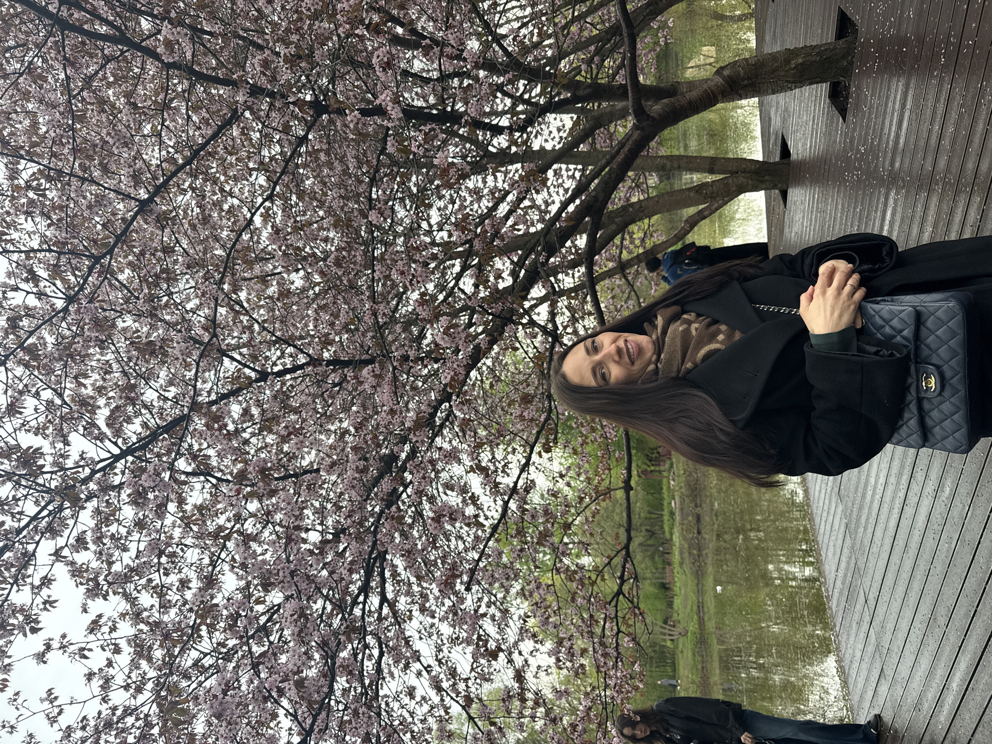 A woman with long brown hair poses under a blossoming tree in Saint Petersburg, Russia. She wears a black coat and a patterned scarf and carries a navy blue quilted handbag. The setting appears to be a wooden walkway near a body of water with other people present in the background.