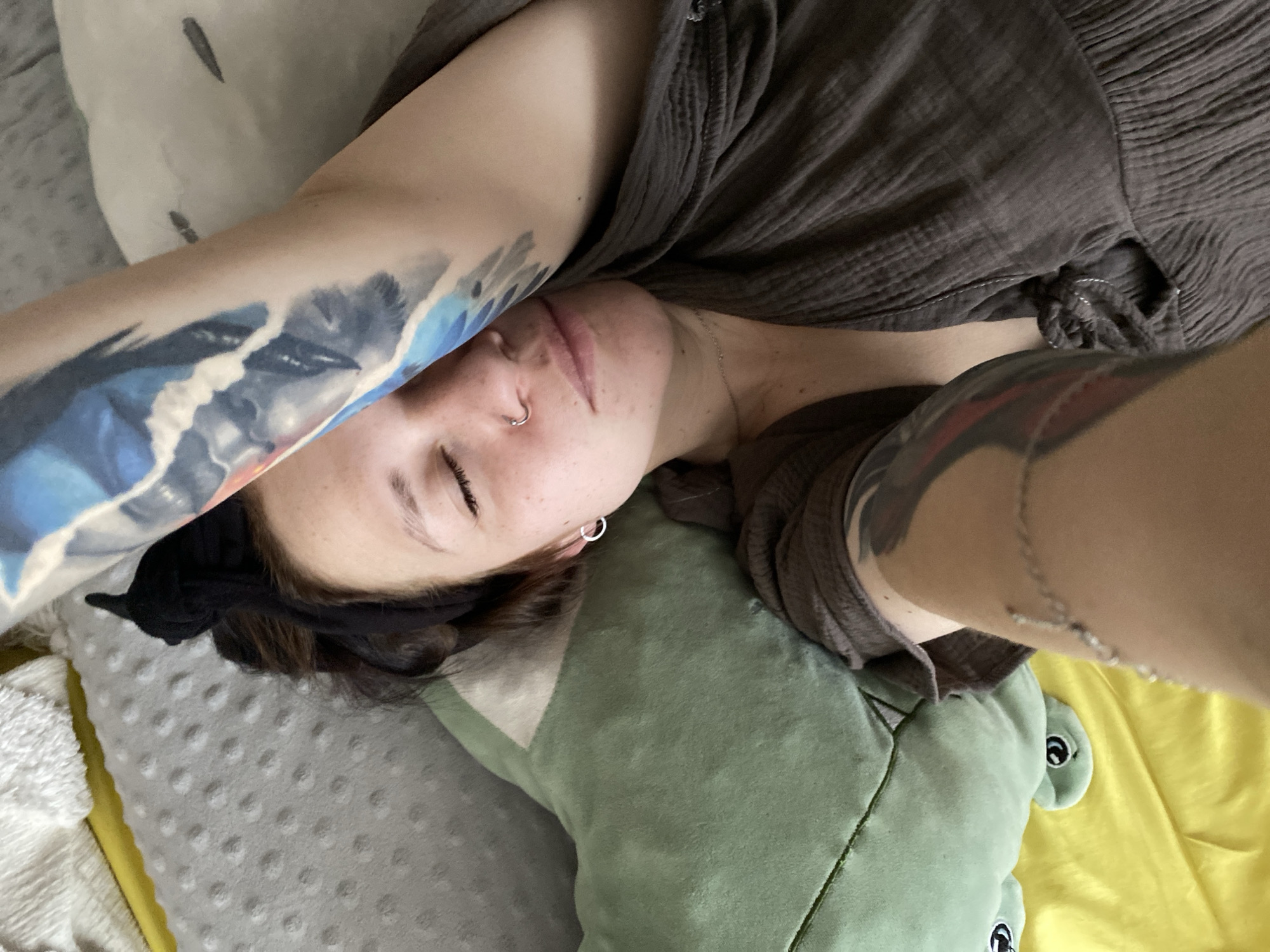 A woman with tattoos on both arms is lying on a bed or couch. She has a nose ring and hoop earrings. Her head rests on a green stuffed alligator. The bed or couch has a gray, dotted blanket and a yellow blanket. She wears a brown shirt and a black headband. The location is not discernible from the image. The city of Chaikovsky, Russia, cannot be determined from the visual information provided.