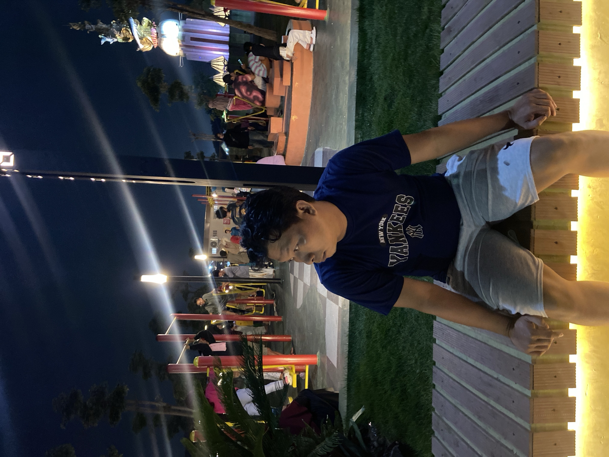 This is a medium shot, outdoor, nighttime photograph taken at Alun-Alun Sidoarjo in Sidoarjo, Indonesia. The central subject is a man with dark hair and skin, wearing a navy blue t-shirt with the New York Yankees logo and light-colored shorts. He is seated on a wooden bench with glowing yellow lights underneath, looking to his right. In the background, various people are visible engaging in activities at a park-like setting. Some individuals are using outdoor exercise equipment with red and yellow frames. Others are gathered around a circular, tiered structure, and one person is seen looking at a mobile phone. To the right of the image, a tall, decorative structure illuminated with pink and purple lights stands against the dark sky, with trees and other lights visible in the distance. The ground is covered in grass and a checkered tiled area. The overall atmosphere suggests a public gathering space at night, possibly a tourist attraction or community park.