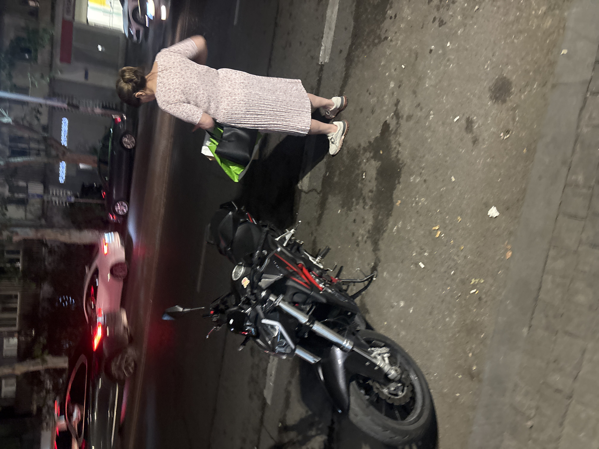 This is a night scene captured on a city street, likely in Tbilisi, Georgia, given the presence of street signs and building architecture consistent with the region. The foreground is dominated by a parked black motorcycle. The motorcycle is sleek, with red accents on its frame and what appears to be a dark-colored seat. Its front wheel is clearly visible, with a disc brake and caliper.
To the right of the motorcycle, a woman is standing, facing away from the camera. She is wearing a light-colored, possibly pink, pleated dress and white athletic shoes. She is holding a large, dark tote bag in her left hand and a bright green shopping bag in her right hand, with its contents partially visible. Her hair is pulled back in a bun.
In the background, several cars are on the road. To the left, a dark-colored car with illuminated taillights is in motion. Further down the street, a white car is also visible. Buildings line the street, with some illuminated windows suggesting activity within. The street itself appears wet in places, and there are scattered cigarette butts on the asphalt. The lighting suggests it is nighttime, with streetlights and car headlights illuminating the scene.