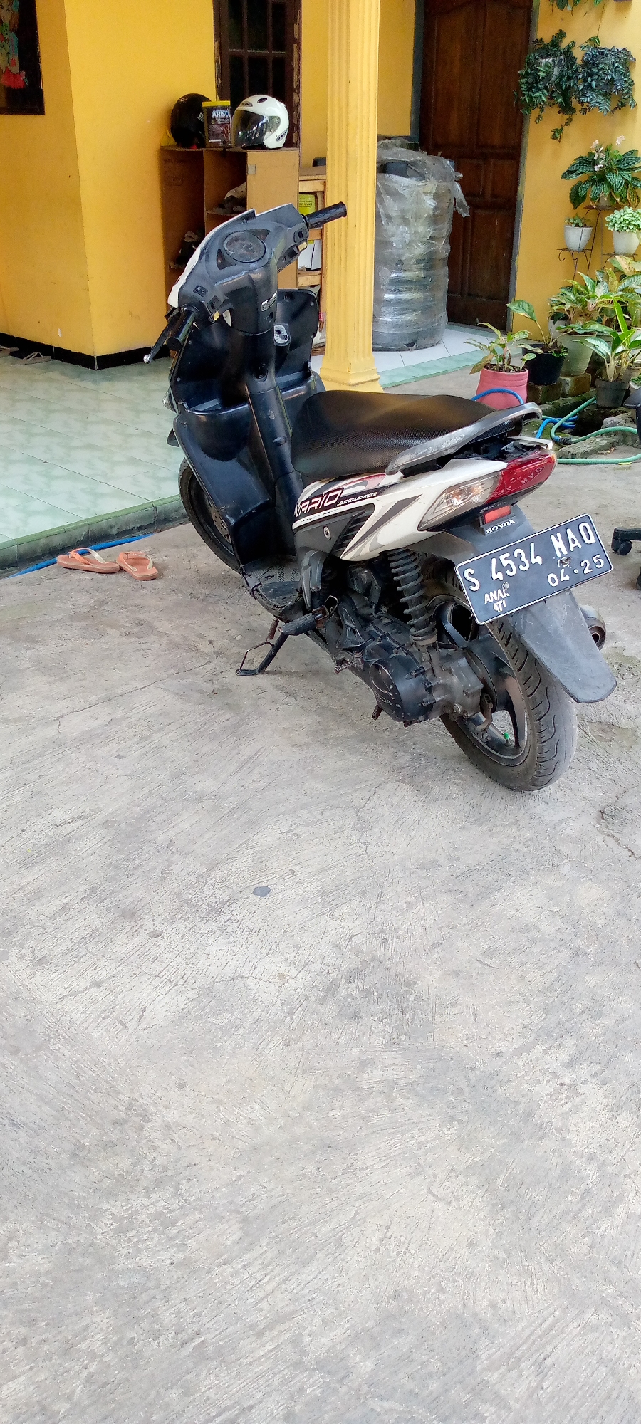 In Sumberwono, Indonesia, a black and white scooter with license plate S 4534 NAQ, dated 04-25, is parked on a concrete surface outside a building with a yellow facade. The scooter is positioned diagonally, facing towards the right of the frame. On a shelf to the left, two helmets, one black and one white, are visible, along with a box labeled "ARISCO". Two pairs of flip-flops are placed on the tiled ground to the left of the scooter. The background reveals a wooden door, a yellow pillar, and several potted plants with green foliage and small white flowers displayed on a tiered stand. A large object wrapped in clear plastic is also visible behind the scooter, near the yellow pillar. The concrete ground is cracked and weathered, indicating an outdoor setting.