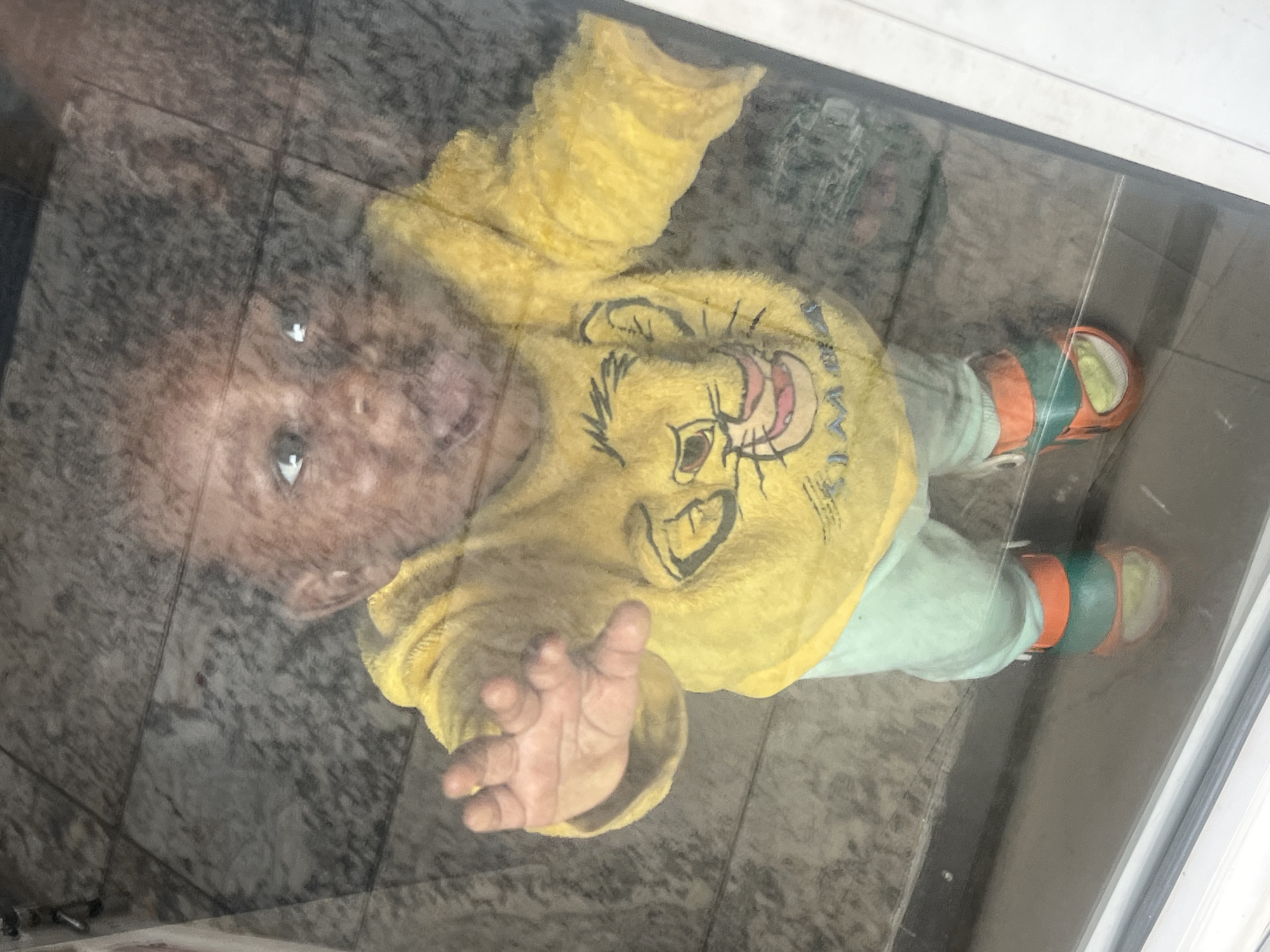 A baby is lying on a glass surface, possibly a tabletop or window, in an undefined location. The baby is wearing a yellow sweatshirt with a Simba print on the front, light green pants, and striped orange and green shoes. The baby's face is visible through the glass, and one hand is outstretched. The background is a patterned gray surface.