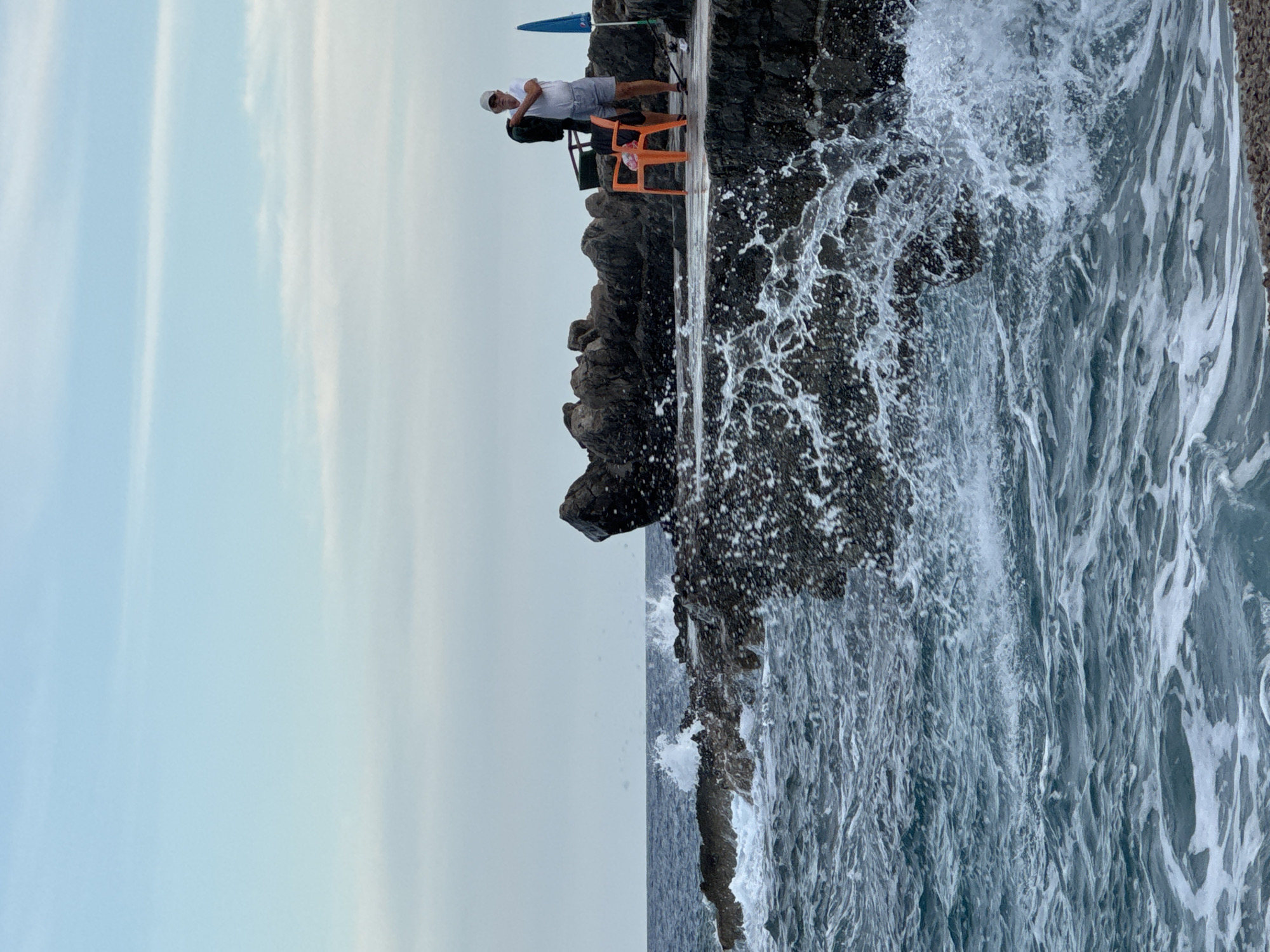 A man stands on a concrete pier next to a rocky outcrop, near the city of Prijevor in Montenegro. He is wearing a white t-shirt, grey shorts, and a white cap, and is holding a dark garment. An orange plastic chair and a green basket are nearby, along with a blue umbrella. Waves crash against the rocks, creating white spray. The sky is overcast with a pale blue hue.