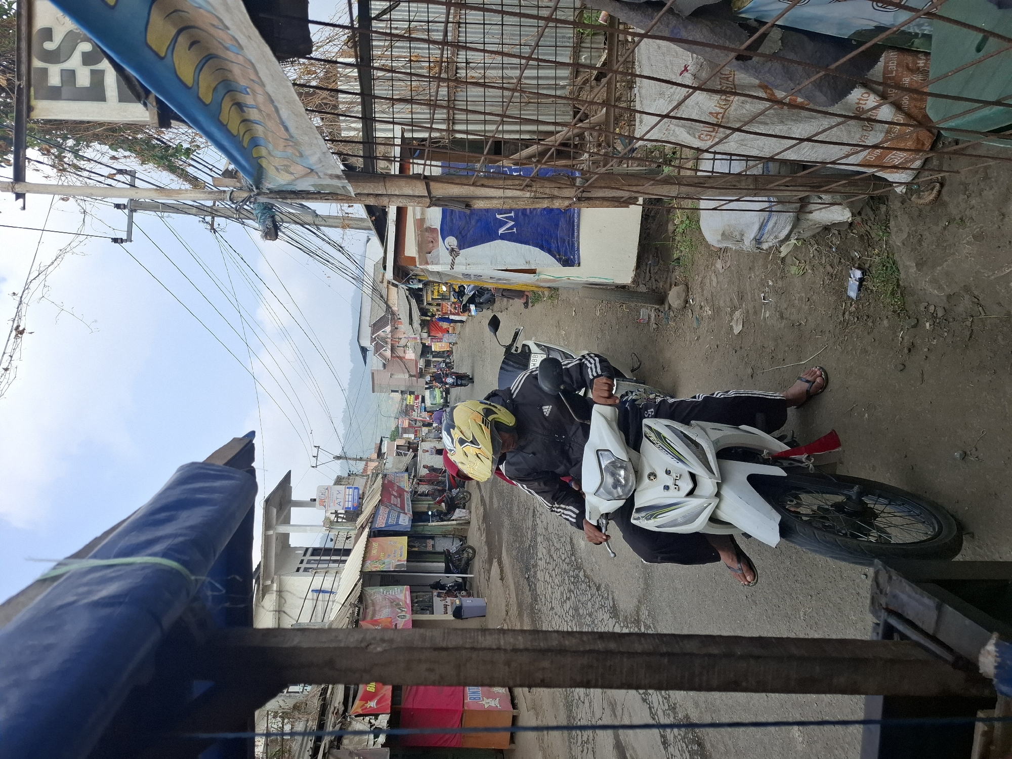 A man wearing a yellow helmet and a black jacket with white stripes is riding a white motorcycle down a street in Ciheulang Tonggoh, Indonesia. The street is lined with buildings and shops, some of which have signs in Indonesian. There are also power lines and trees visible in the background. The man is wearing black pants and flip-flops. The motorcycle has a license plate that is partially visible. The sky is overcast.