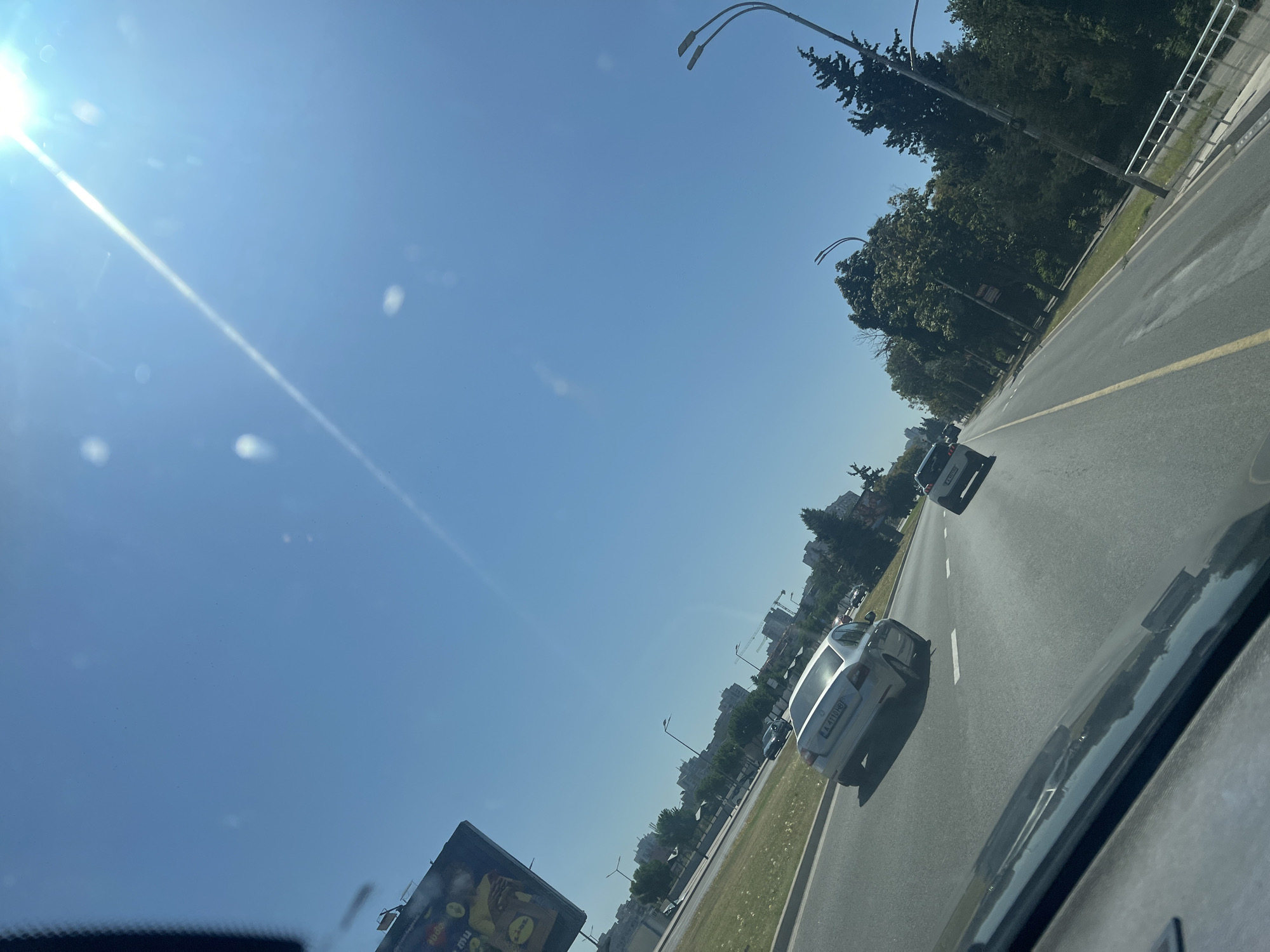 The image captures a view from inside a vehicle, looking out at a sunny day in Burgas, Bulgaria. The road ahead is paved with asphalt, marked with a yellow line down the center and white dashed lines to delineate lanes. Several cars are visible on the road, with the closest being a silver sedan. In the distance, apartment buildings and trees line the side of the road, suggesting an urban or suburban environment. The sky is a clear, bright blue, with the sun prominently positioned in the upper right corner, casting a lens flare across the scene. A large billboard is partially visible on the left side of the frame, advertising an unknown product. The overall impression is of a typical drive through the city on a warm, clear day.