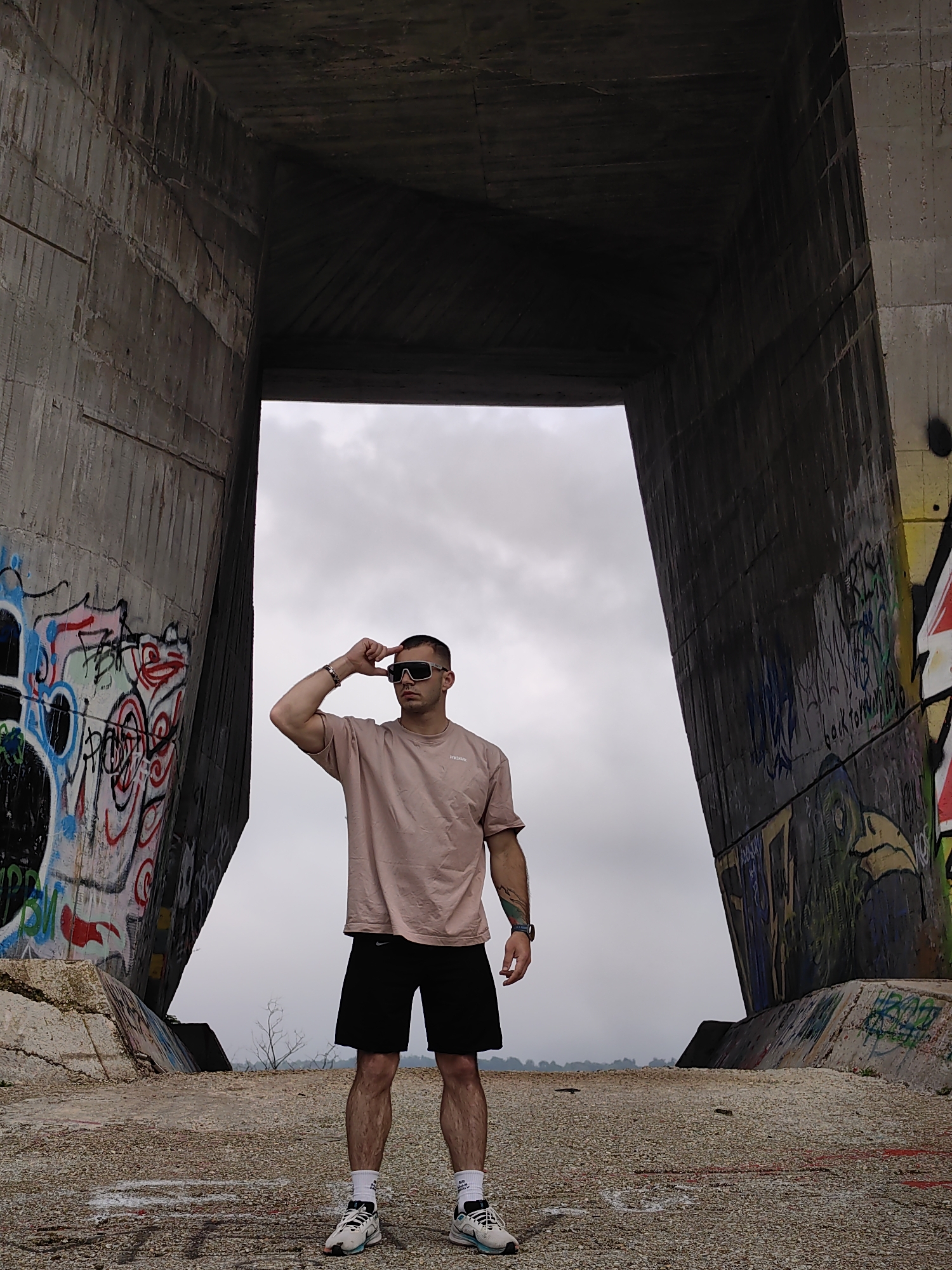 A man stands in the center of the frame under a large, open, concrete structure in Varna, Bulgaria. He is wearing a light pink t-shirt, black shorts, white athletic socks, and athletic shoes. He is also wearing dark sunglasses and has a tattoo on his left forearm. He is touching the rim of his sunglasses with his right hand. The concrete structure, likely a bridge or architectural feature, has graffiti on its inner walls. The ground is rough and uneven. The sky is overcast.