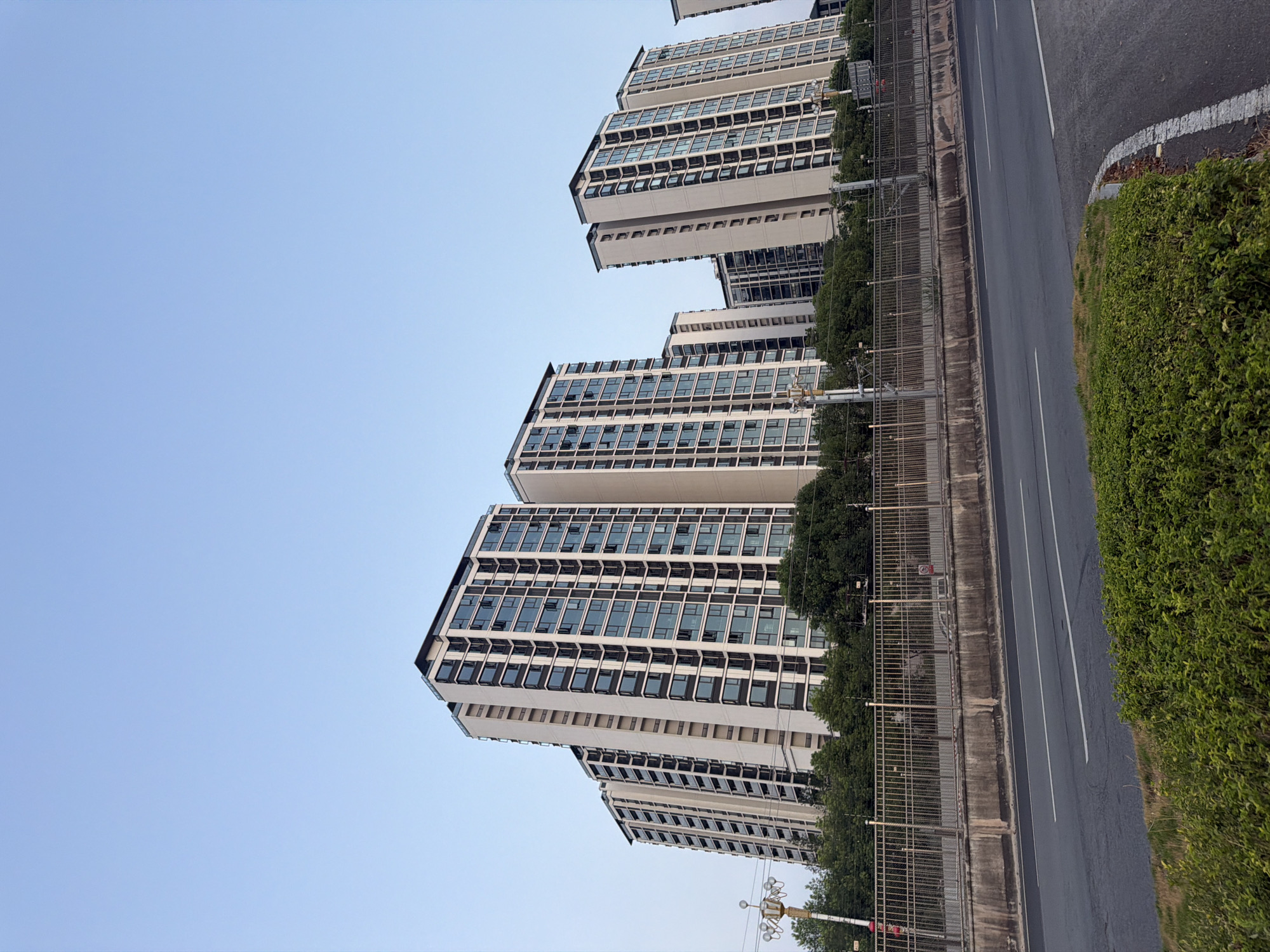 The image shows a view of apartment buildings in Jiangjin, China. In the foreground, there is a road with white lane markings, bordered by a strip of green grass and bushes. Beyond the road, a metal fence separates it from a concrete barrier and a grassy embankment. Behind the fence, a line of trees forms a green canopy, and further back, several tall apartment buildings are visible. The buildings are modern, with many windows and balconies. The sky above is a clear, pale blue, indicating it is likely daytime. There are no people visible in the image.