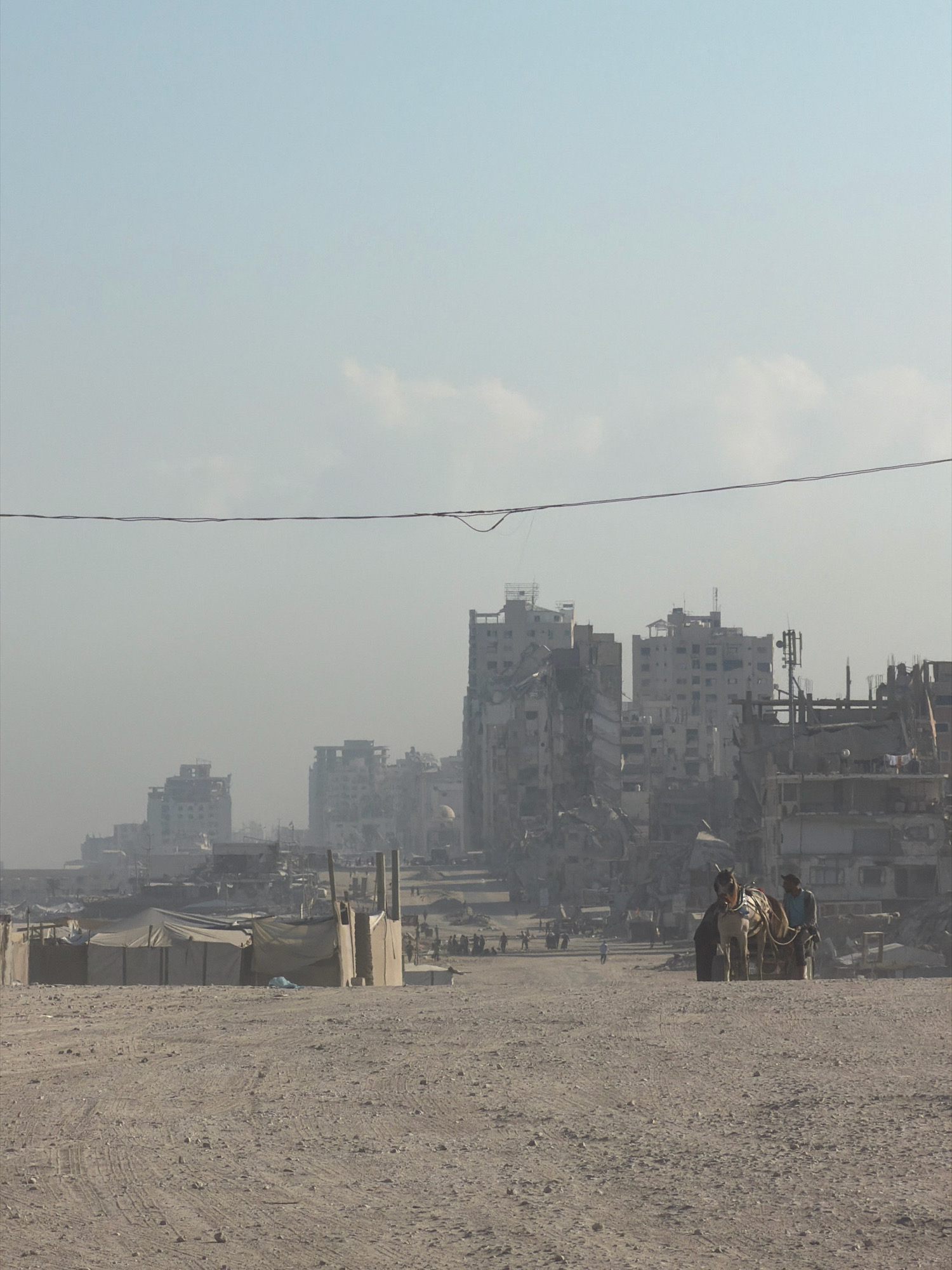 An outdoor, eye-level, medium shot captures a desolate and war-torn urban landscape in Gaza under a hazy sky. In the foreground, a barren, sandy terrain is marked with tire tracks and scattered debris. A makeshift camp of tents and structures, constructed from canvas and wood, stands in the mid-ground to the left.
Further into the scene, a horse-drawn cart, occupied by two individuals, moves across the open area. The cart is laden with goods, and the individuals appear to be navigating the rough terrain.
The background is dominated by the skeletal remains of bombed-out buildings, their damaged structures and exposed interiors indicative of extensive destruction. In the distance, more intact buildings suggest a city that has been partially rebuilt or spared from the worst of the damage. The overall atmosphere is one of ruin and resilience, with signs of life persisting amidst the devastation.