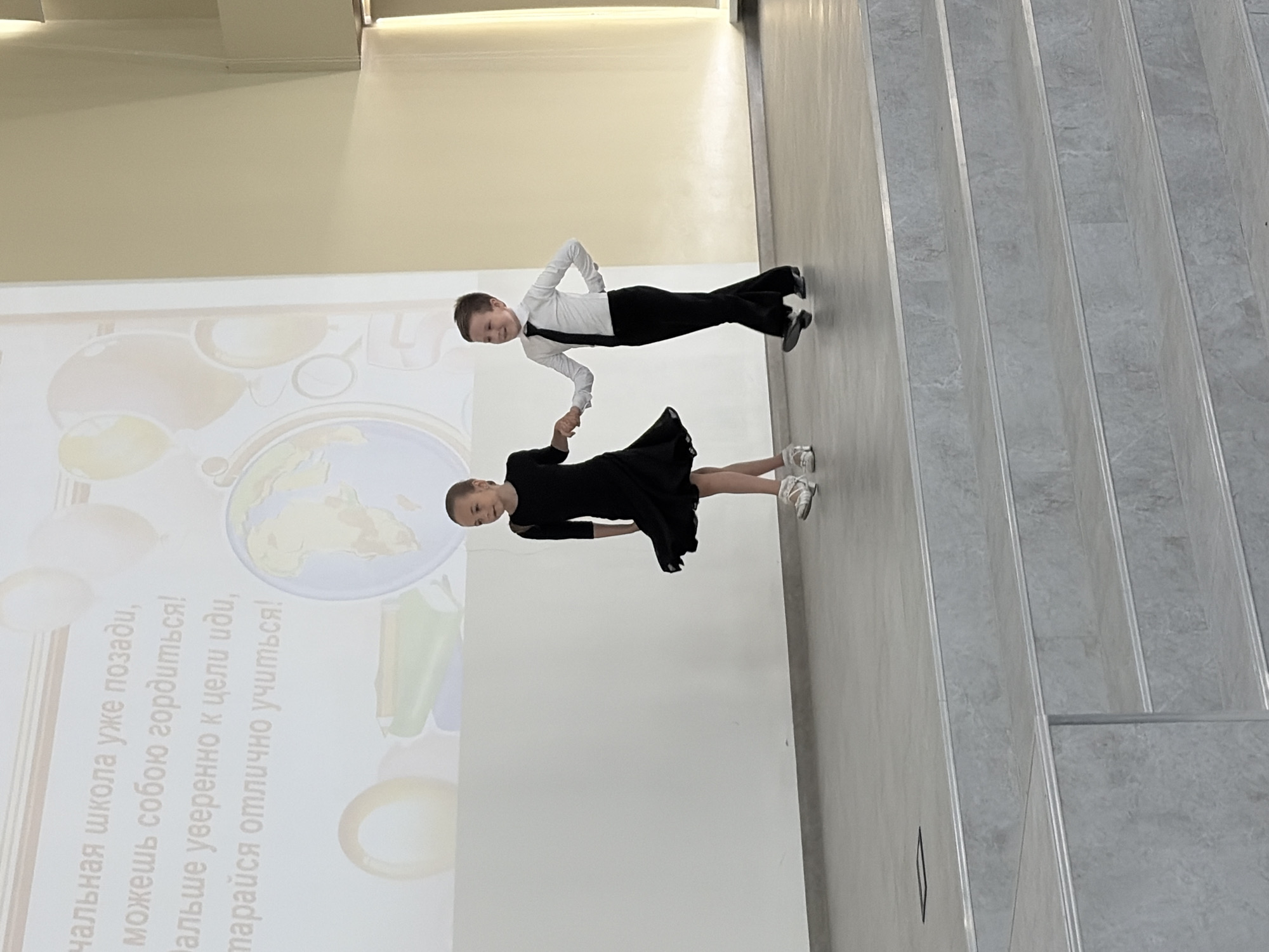 Two young children, a boy and a girl, are captured in a dance pose in what appears to be an indoor setting. The boy is wearing a white shirt, black pants, and a black tie, while the girl is in a black dress. They are holding hands, suggesting they are dance partners. The background features a wall with a projection displaying text in Russian, along with illustrations including a globe. Adjacent to the wall is a staircase with gray steps. The overall location is identified as Chaikovsky, Russia.
