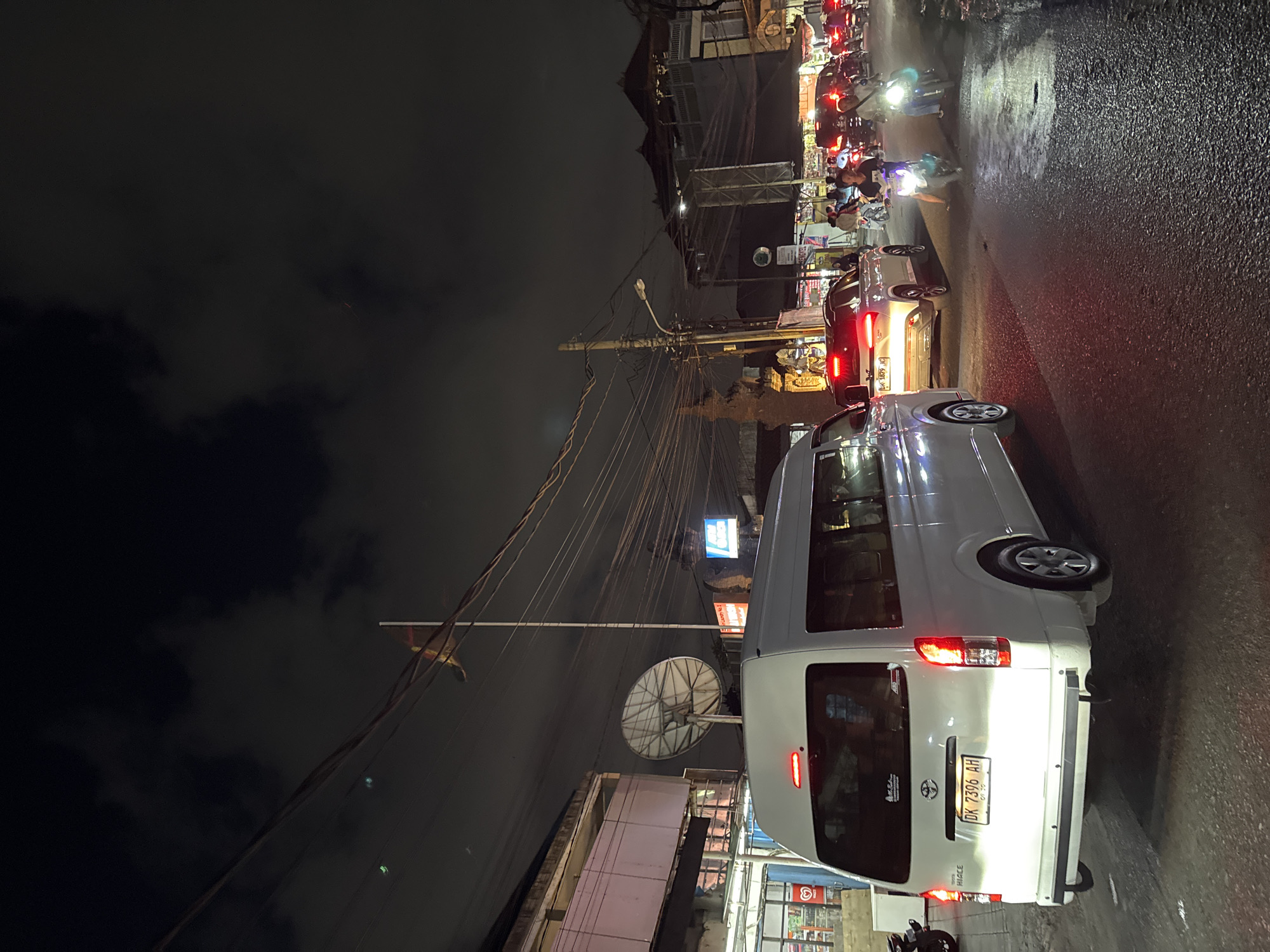 This is a nighttime, eye-level, medium shot of a busy street in Denpasar, Indonesia. The image is dominated by a white van in the foreground, its rear lights illuminated. To the right of the van, a white SUV is also visible with its brake lights on. The road is wet, reflecting the lights of the vehicles and streetlights. Several motorcycles are present, some with riders, contributing to the traffic. The background shows buildings with some illuminated signs and a dense network of overhead electrical wires. The sky is dark and cloudy, with hints of moonlight. The overall impression is one of a typical bustling street scene at night in a Southeast Asian city.