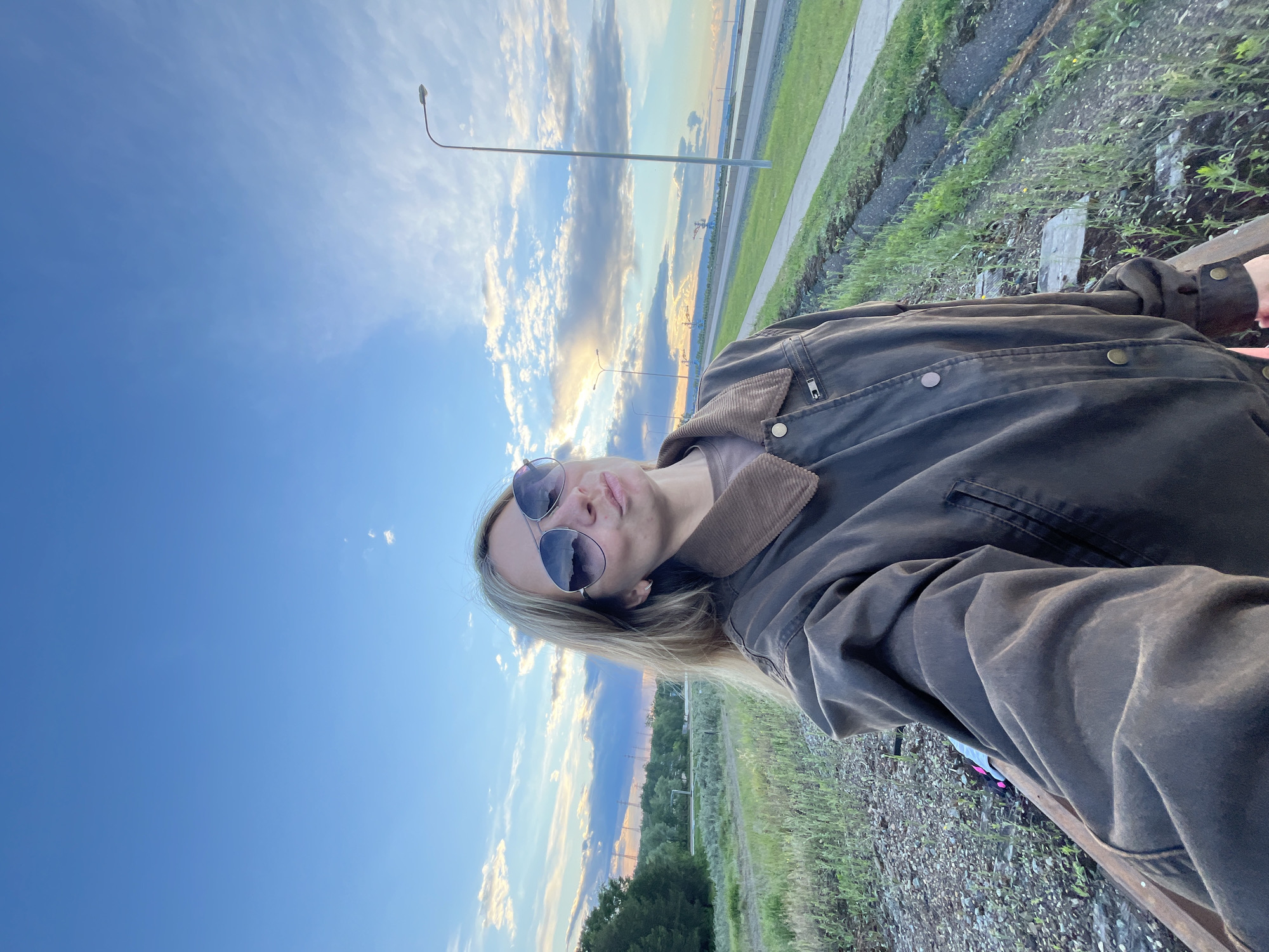 A young woman wearing sunglasses and a brown jacket takes a selfie outdoors. The location appears to be near a road in the city of Chaikovsky, Russia, as indicated by the presence of a street lamp and the cityscape in the background. The sky is blue with some clouds. The woman is sitting near vegetation alongside the road.