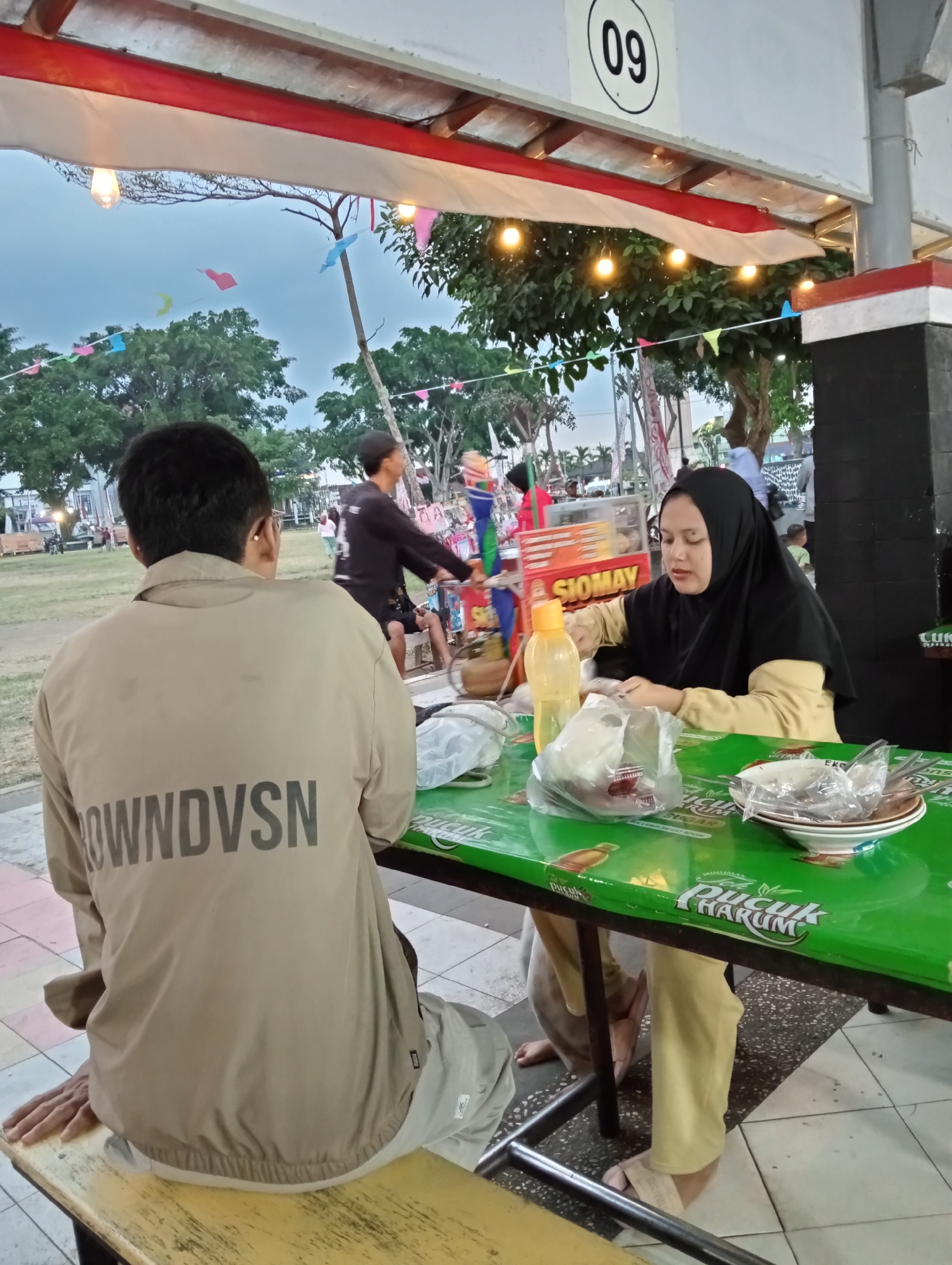 The image depicts a casual outdoor dining scene at a stall in Magelang, Indonesia. Two individuals are seated at a bright green table. In the foreground, a person wearing a tan jacket with "ROWNDVSN" printed on the back is seen from behind. To the right, a woman wearing a black hijab and a yellow shirt is actively engaged with food on the table, which includes plastic-wrapped items and a bottle of liquid. The table itself is adorned with various logos, including "PUCK HARUM". In the background, a food stall with the word "SIOMAY" prominently displayed is visible. The environment suggests a lively atmosphere, with trees, decorative flags, and streetlights in the background, hinting at a public area or park, potentially a night market or a similar gathering spot within the city of Magelang.