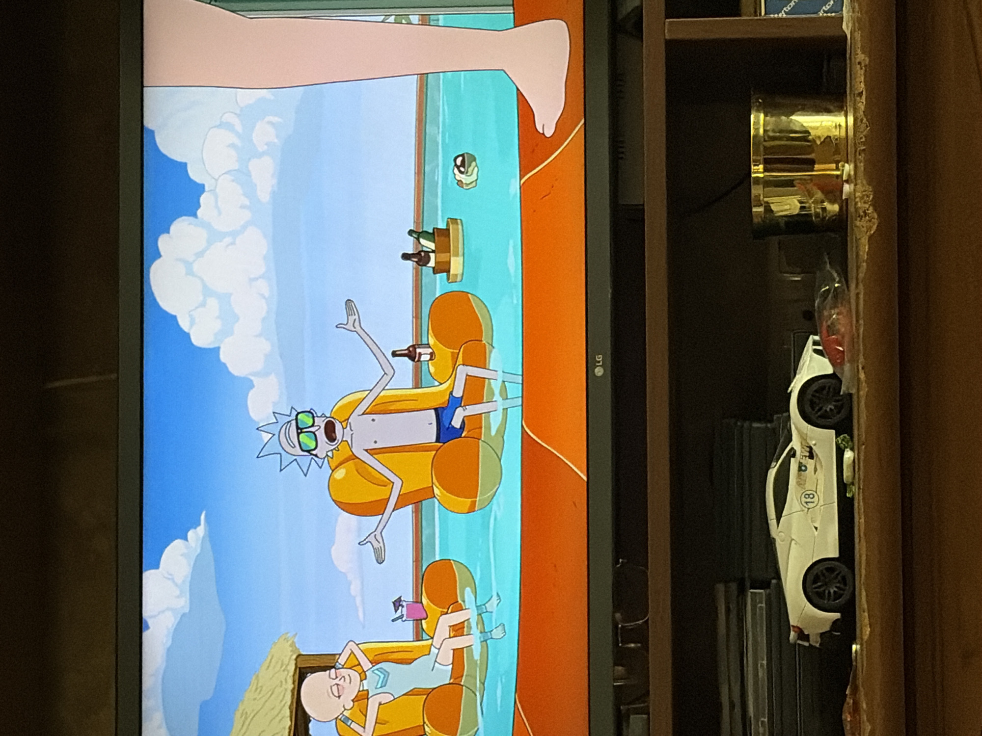 In an animated scene, two bald, humanoid figures are relaxing on inflatable chairs in a swimming pool under a bright blue sky with fluffy white clouds. The figure on the left, wearing a light blue swimsuit, reclines with their arms behind their head. The figure on the right, wearing blue swim trunks, has their arms outstretched and mouths open as if speaking. There are drinks and bottles floating in the pool. To the right, a large, light-skinned leg is visible, suggesting another person is present. The scene appears to be set in a resort or a vacation location. Below the screen, on a wooden shelf, rests a white toy sports car with the number 18 on its side, and a golden cylindrical container.