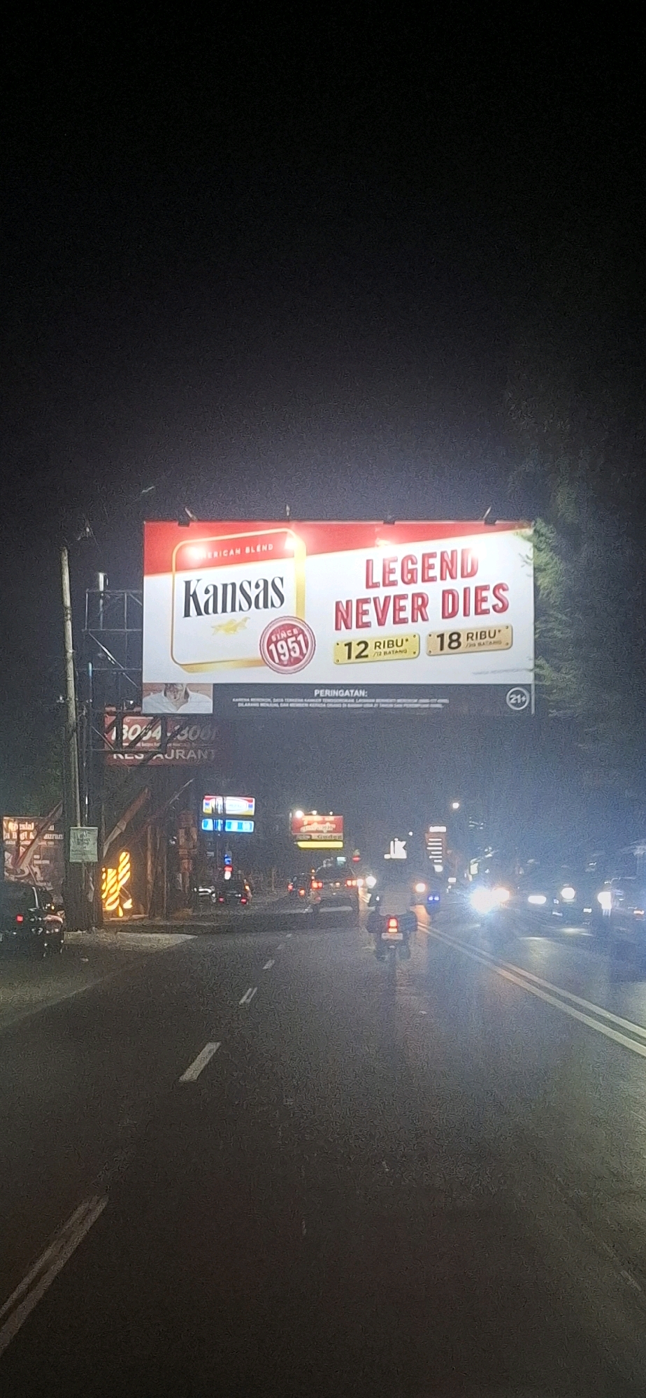 An outdoor, eye-level, vertical shot captures a billboard advertising Kansas cigarettes at night on a street in Colomadu, Indonesia. The billboard features a red and white design with the brand name "Kansas" prominently displayed, along with the tagline "LEGEND NEVER DIES" and pricing information. Below the billboard, a sign for "Restaurant" is visible. The street is paved and illuminated by streetlights and the headlights of passing vehicles, including cars and motorcycles. The scene depicts a typical urban night setting with traffic and commercial signage.