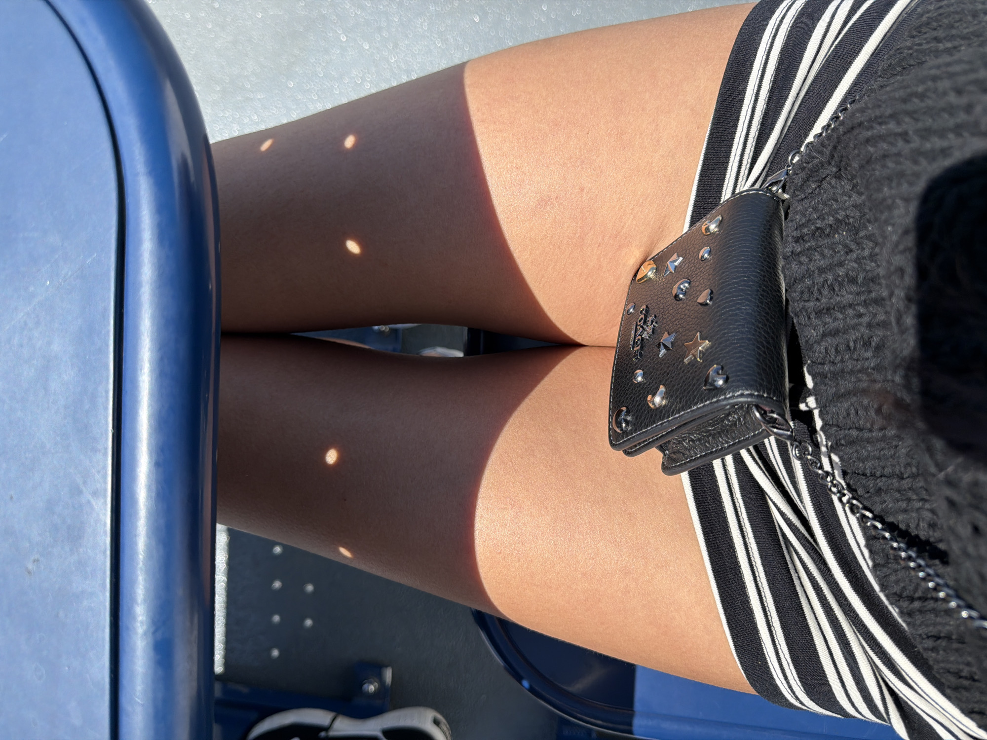 The image shows a close-up view of a person's legs and a wallet. The person is sitting on a blue seat, as evidenced by the blue structure at the top of the image and the blue seating visible on the left. Sunlight is filtering through, creating dappled shadows on the person's legs. The person is wearing a black and white striped skirt or shorts, and a black knitted top. A small black wallet, adorned with silver studs and stars, is placed on the person's lap. The background appears to be a textured surface, possibly a wall or ground, with small, bright circular reflections. The scene is shot from a high angle, looking down at the person's legs. There is no identifiable information about the location being in Southern Gulf Islands Electoral Area, Canada within the image itself.
