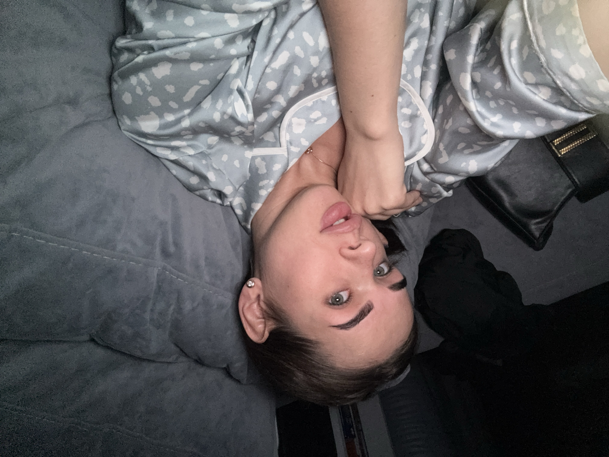 The image shows a woman lying on a gray, textured surface, possibly a couch or bed, wearing a light blue and white patterned pajama top. Her head is positioned upside down in the frame, and her hair is pulled back. She is looking directly at the camera. Her right hand is near her neck, and she is wearing a thin necklace. To the right of the frame, there is a black object, possibly a purse or bag. The background is dark. There is no evidence to suggest the location is Лобаново, Russia.
