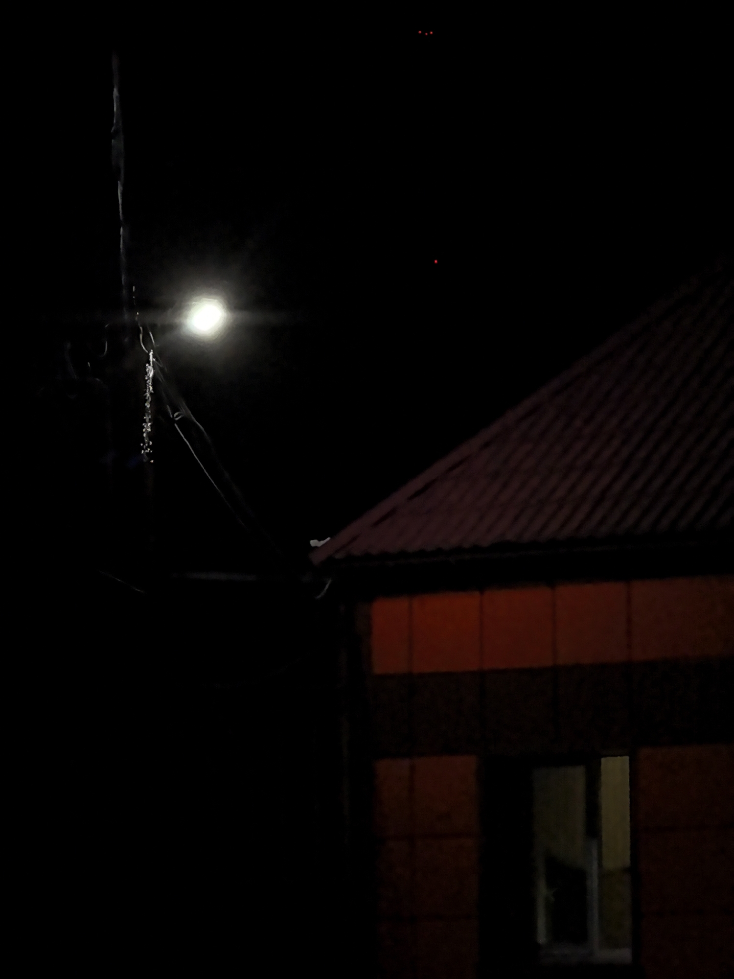 An outdoor nighttime scene shows a street lamp illuminating a dark sky and a portion of a building. A single bright light source from the street lamp is visible to the left of the frame. To the right, a building with a dark-colored, sloped roof is partially visible. The building's facade features horizontal bands of red and dark material. A dark window with a visible frame is located in the lower right section of the building. In the upper portion of the black sky, three small red dots are discernible, two clustered together and one slightly below and to the left. The environment appears to be urban or suburban.