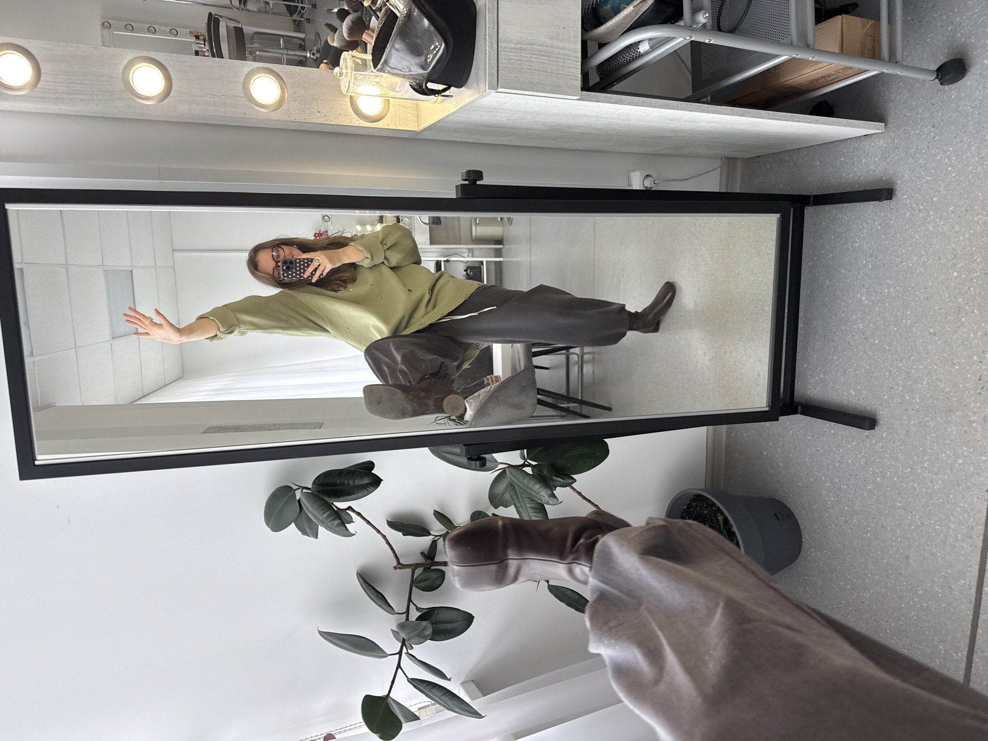 A person wearing glasses and a mask takes a selfie in a mirror with a black frame. The person is wearing a green sweater and dark pants and is extending their left arm. The mirror is next to a potted plant with dark green leaves. A row of lights is visible at the top of the mirror frame, and storage shelves are visible in the background. The wall behind the mirror is white. The location cannot be determined from the image.