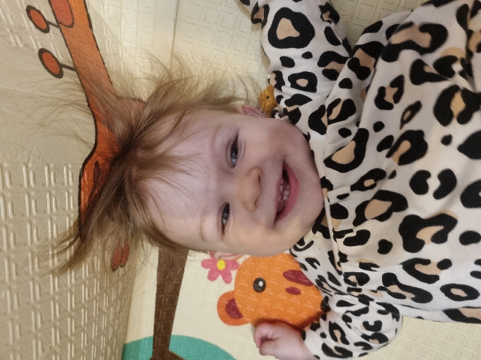 A baby lies on a patterned playmat, wearing a white onesie with black and brown leopard spots. The baby smiles, showing teeth. Patches of orange with a cartoon bear head are visible on the mat. The baby's hair is tousled, and blue eyes are visible. A drawn giraffe neck is also on the mat.