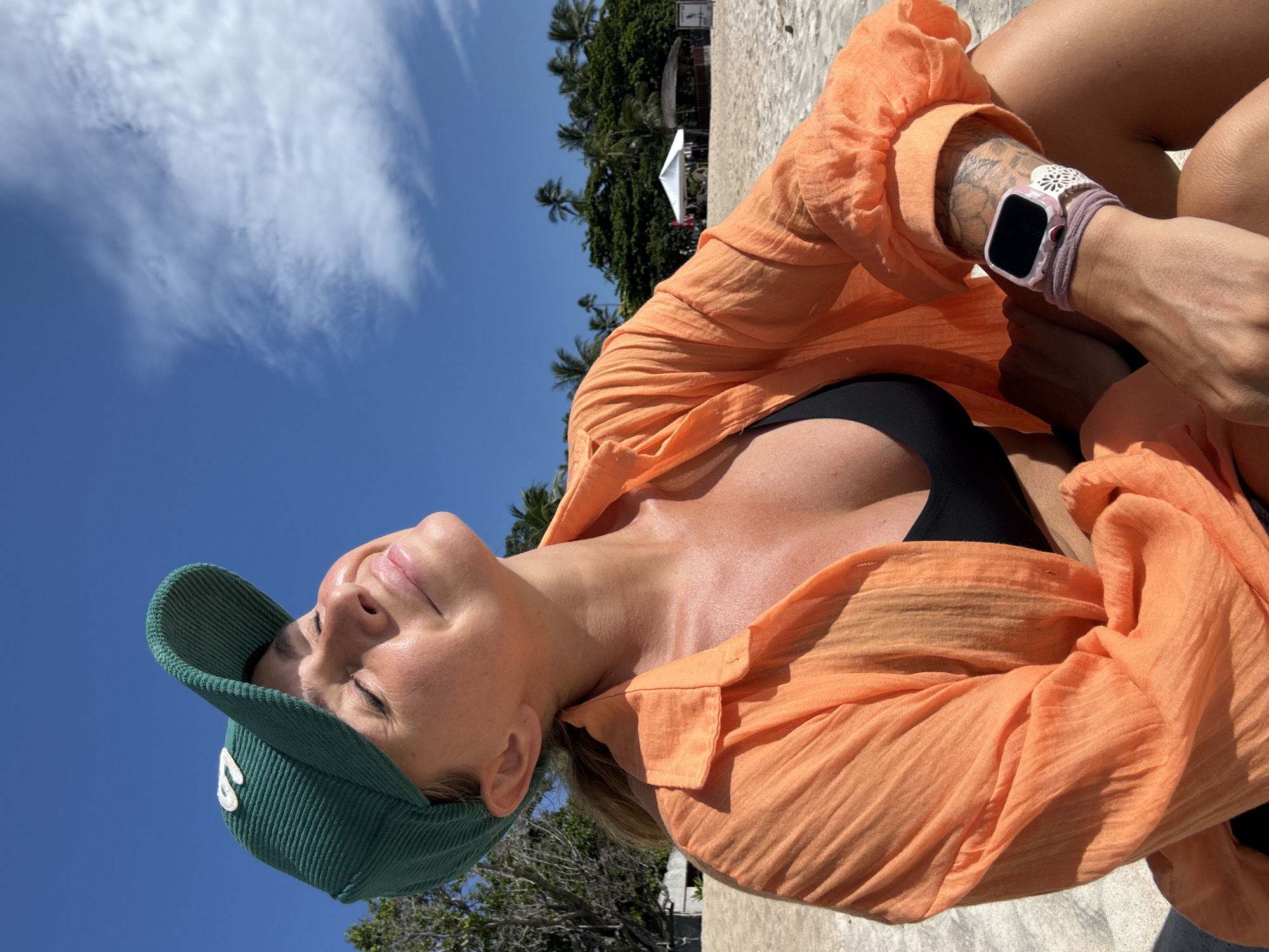 A woman is sitting on a sandy beach in Ko Samui District, Thailand, with her eyes closed and face tilted towards the sun. She is wearing a green corduroy baseball cap, an orange unbuttoned shirt, and a black bikini top. A smartwatch with a pink and white patterned band is visible on her left wrist. Her right arm rests on her lap, revealing a tattoo on her forearm. In the background, palm trees and a white tent can be seen on the beach, under a bright blue sky with scattered clouds.