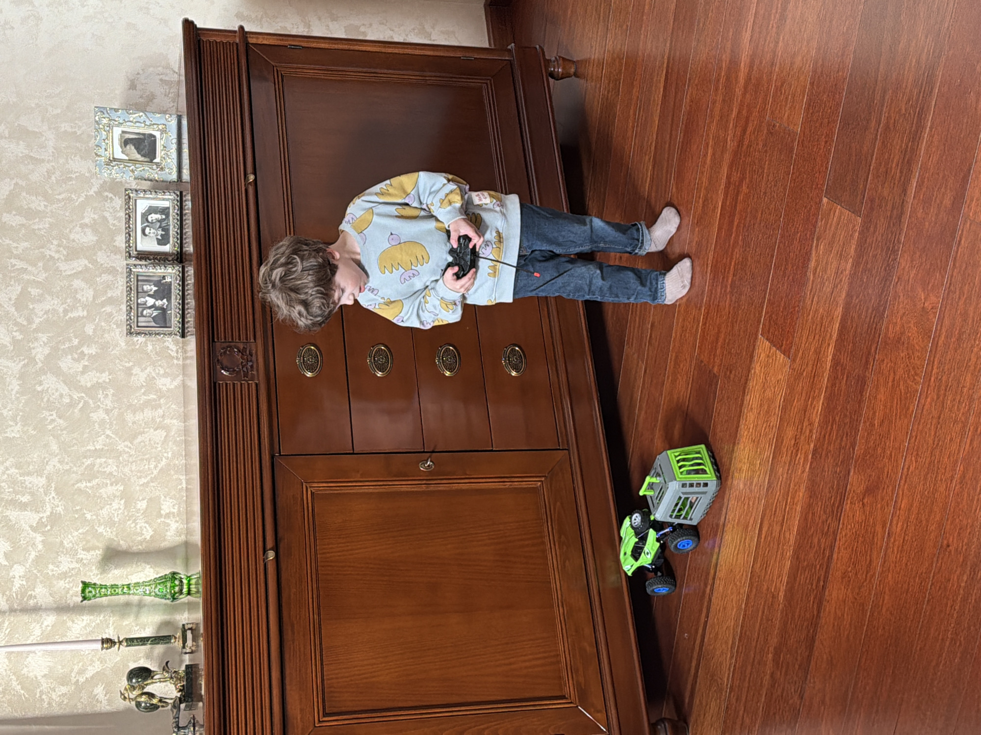 A boy stands in socks on a polished hardwood floor controlling a green and grey toy truck with a remote. He is wearing jeans and a light blue sweatshirt with yellow bird patterns. The floor is next to a large, dark wood cabinet or wardrobe with metal drawer pulls. To the left of the cabinet, mounted on the wall, are three framed photos and a decorative glass vase and metal decorative object. The scene appears to be indoors. There is no information available to ascertain whether this image was taken in the city of Mishutkino, Russia.