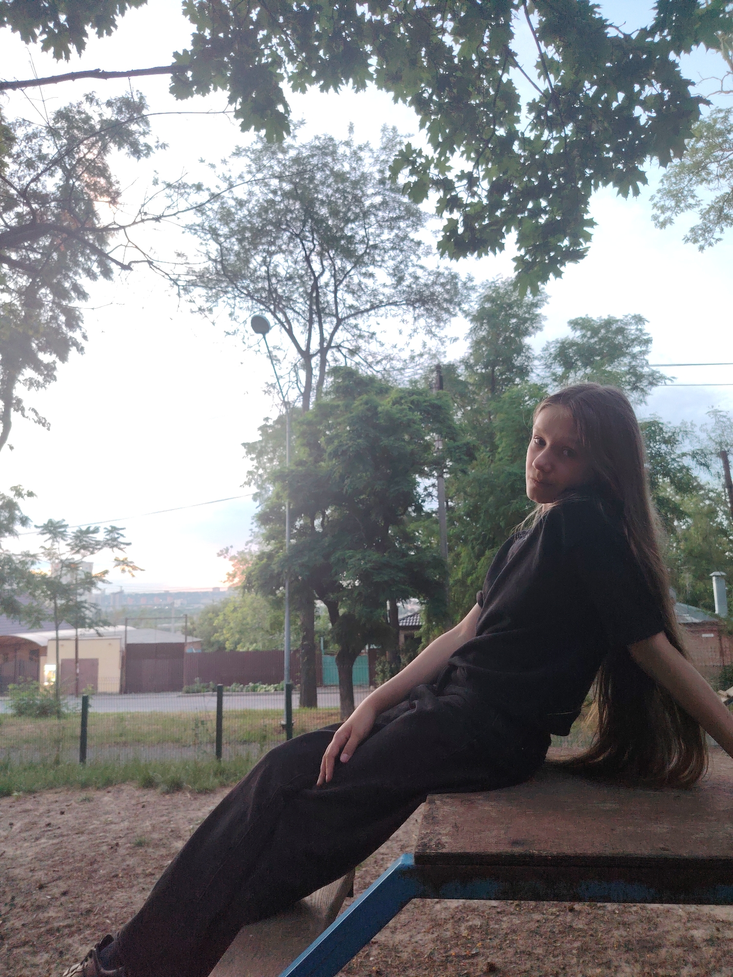 The image depicts a young female with long, dark hair sitting outdoors on a wooden structure with blue metal legs. She is wearing a black t-shirt and black pants. Her body is angled towards the right, and she is looking back towards the camera with a neutral expression. The background shows a park-like setting with trees and a street with a fence and buildings in the distance. The sky is overcast.
This scene is likely located in Rostov-on-Don, Russia, given the style of the buildings and vegetation, which are consistent with urban parks and residential areas in that region. The presence of trees, grass, and a playground-like structure suggests a public outdoor space. The lighting indicates it is likely daytime, possibly late afternoon or early evening due to the soft light. No specific landmarks identifiable to Rostov-on-Don are visible, but the overall aesthetic is plausible for the city.