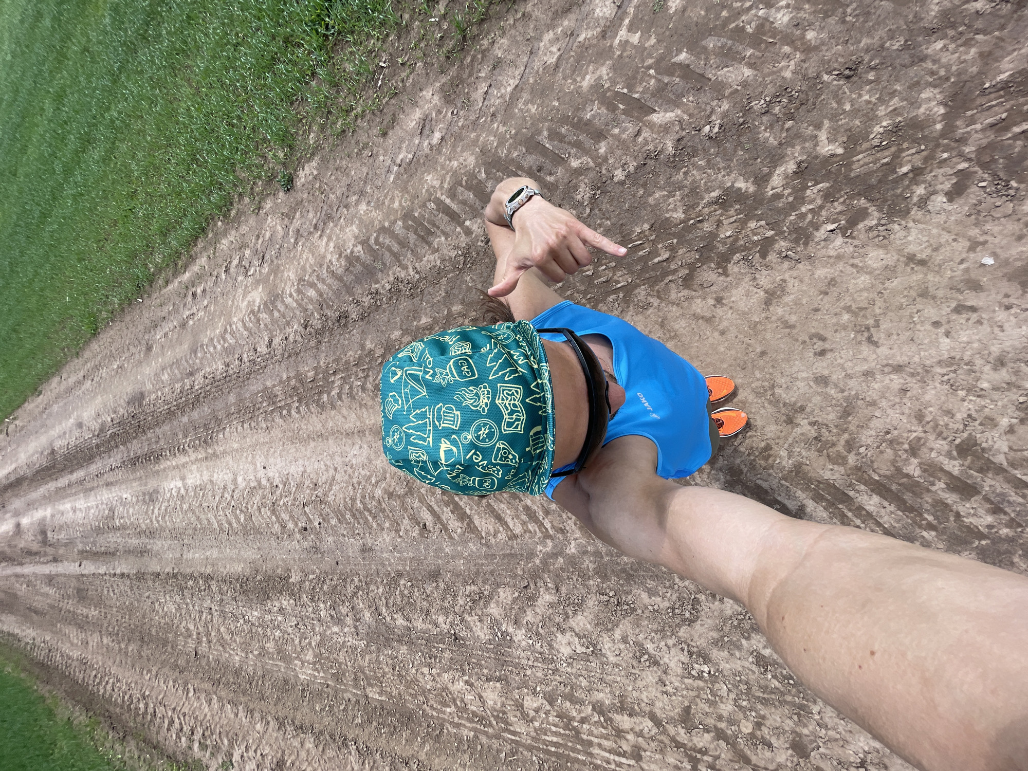 The image depicts a first-person, overhead perspective of an individual standing on a dirt road, likely taken as a selfie. The location is an outdoor, rural or semi-rural setting in Unknown, Unknown city.
In the foreground and center, the upper torso and head of a person are visible. The person is wearing a teal-green cap patterned with various white graphics, including trees, a compass, a campfire, a coffee cup, a beer stein, a slice of pizza, a map, mountains, and ambiguous text resembling "240" or "CAD." Dark sunglasses are pushed up onto the cap. Below the cap, a bright blue sleeveless athletic top with a visible "JAKO" logo on the right chest is worn. The person's left hand is extended, making a "shaka" or "hang loose" gesture (thumb and pinky extended, middle fingers curled). A light-colored wristwatch with a dark face is on the left wrist. The person's right arm extends into the immediate foreground, obscuring part of the lower body and likely holding the camera. The individual's feet, clad in bright orange athletic shoes with white soles, are visible near the bottom of the frame.
The ground consists of a dry, light brown dirt road heavily marked with deep, parallel tire tracks, indicative of agricultural or heavy off-road vehicle use. The dirt surface is uneven, with scattered small stones and compacted areas. To the right of the dirt road, a dense field of vibrant green grass extends towards the background. The scene is brightly illuminated, consistent with daylight conditions.