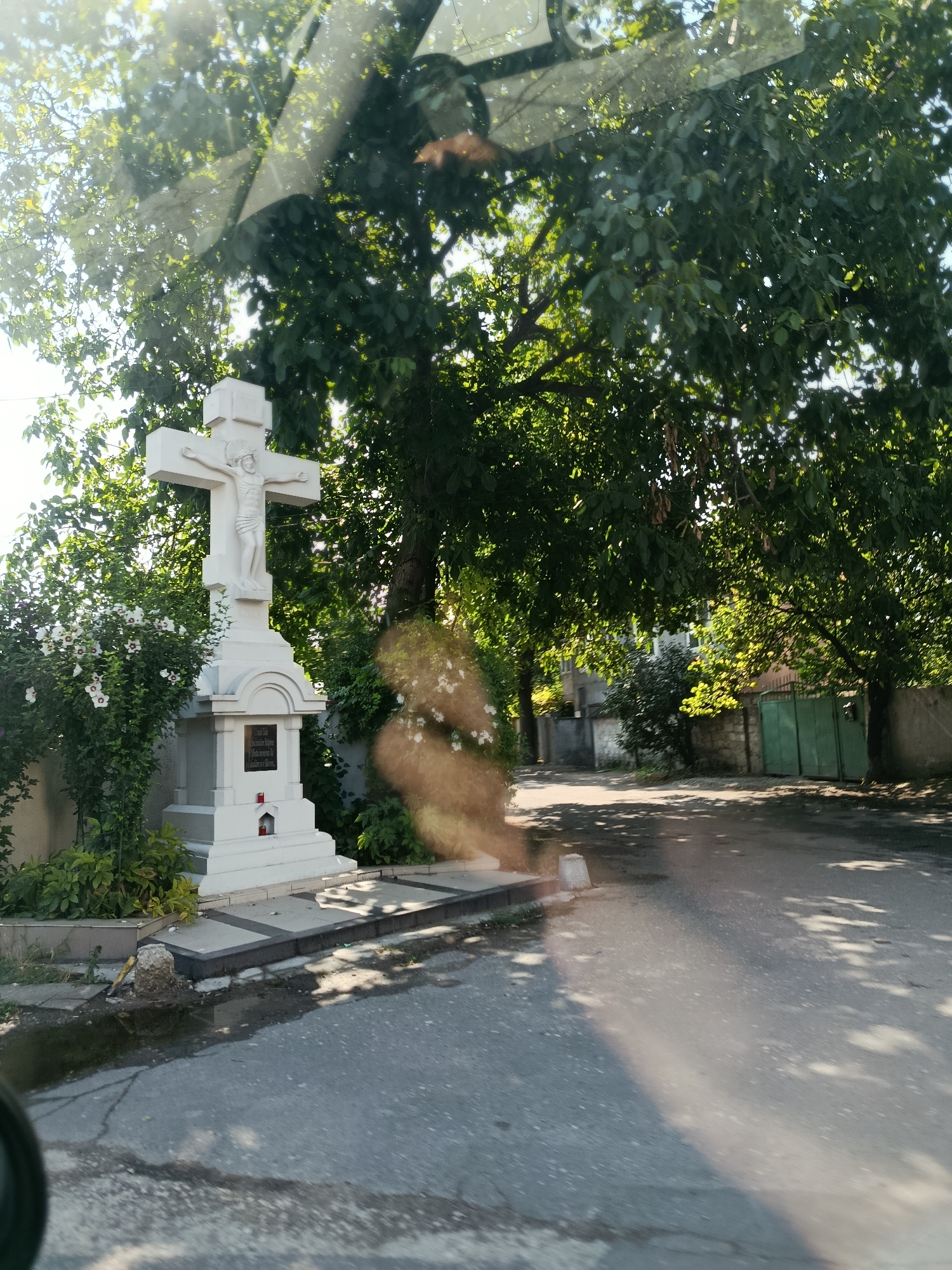 This image captures a white stone crucifix with a depiction of Jesus Christ prominently displayed on a pedestal. The crucifix is situated outdoors, beside a paved road, amidst lush greenery and mature trees casting dappled shadows. A patch of white flowers blooms on a bush to the left of the crucifix. A small red candle is visible within a niche at the base of the pedestal. In the background, a stone wall, a weathered green gate, and residential buildings are discernible. The scene appears to be a roadside shrine in Chișinău, Moldova, given the context provided by the prompt. The overall ambiance is serene and evokes a sense of reverence, with sunlight filtering through the leaves, illuminating the area.