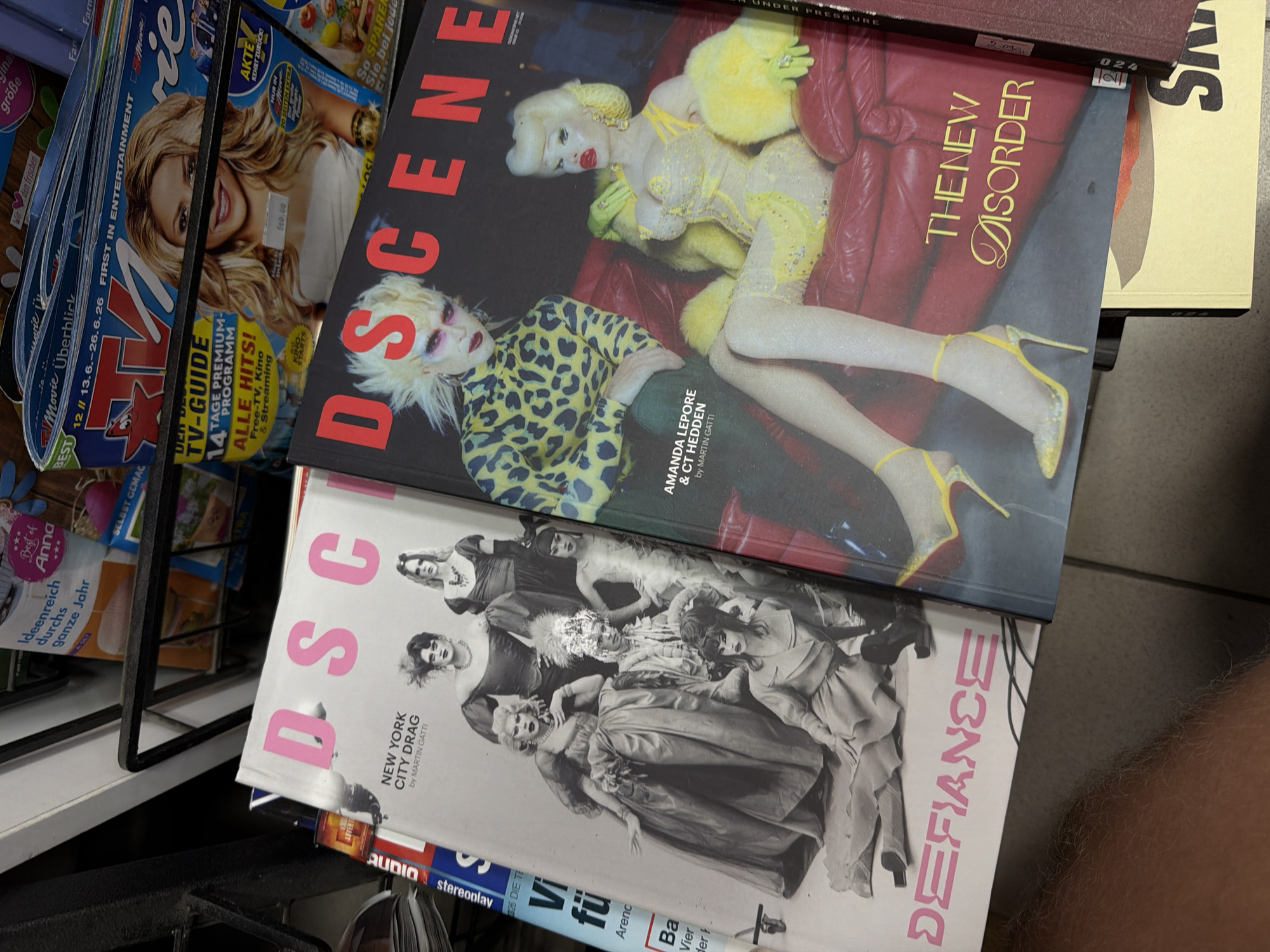 This media file is a photograph of a magazine display, likely in a retail setting. The magazines are stacked and at an angle, with the covers of several prominently visible.
The most prominent magazine in the foreground features a dramatic, stylized photograph of two individuals with elaborate makeup and costumes. One figure, with bright blonde hair and pale skin, wears a yellow, feathered outfit and sits on a red couch. The other figure, also with blonde hair and distinctive makeup, wears a leopard-print top. Text on this cover includes "DSCENE", "AMANDA LEPORE & CT HEDDEN by MARTIN GATTI", and "THE NEW DISORDER".
Below this magazine, another cover is visible, also featuring the title "DSCENE". This cover displays a black and white photograph of a group of people in formal attire, with "NEW YORK CITY DRAG by MARTIN GATTI" and "DEFIANCE" printed on it.
To the left, the covers of other magazines are partially visible. One has a blonde woman with long hair on its cover, and text including "TV-GUIDE", "PREMIUM-PROGRAMM", and "ALLE HITS!". Other magazines show various designs and some German text like "Ideenreich", "gross", and "Frischen".
The setting appears to be indoors, possibly a newsstand or a shop selling magazines and media. The lighting suggests an ambient indoor environment. The overall impression is a vibrant and possibly avant-garde collection of publications.