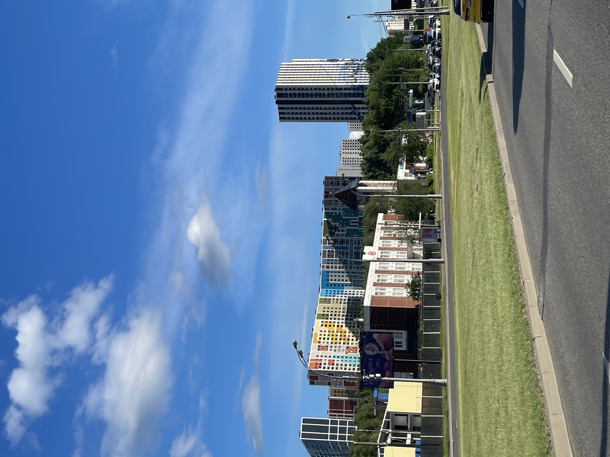 The image shows a cityscape in Moscow, Russia, likely taken from a road running alongside a grassy median strip. Tall, modern apartment buildings with colorful facades are visible on the left, some featuring repetitive square window patterns in vibrant hues like yellow, orange, blue, and white. Further back, taller, more generic buildings are present. The sky is clear blue with scattered puffy white clouds. A portion of a roadway with asphalt and a painted white line are seen in the foreground on the right, where a car is partially visible. Street lamps are also present. The scene is bathed in bright sunlight, suggesting a clear day.