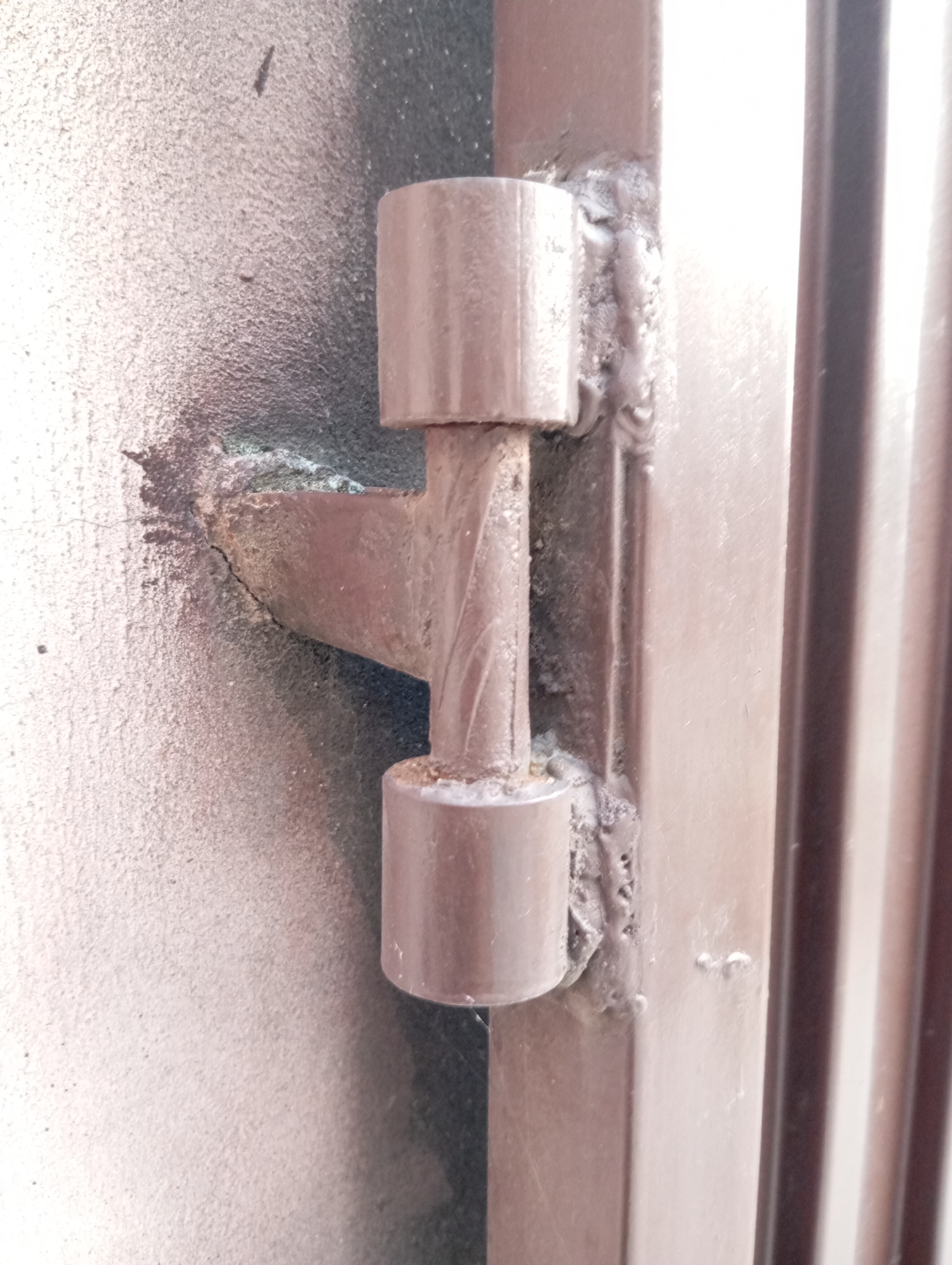 The image shows a close-up, eye-level, medium shot of a metal hinge on what appears to be a gate or door. The hinge is welded onto a vertical metal post, which is part of a gate or door frame. The hinge itself consists of two cylindrical parts, one attached to the frame and the other to the gate, which interlock to allow for rotation. The welding is visible and appears to be crude, with some unevenness and blobs of metal. The surfaces of the hinge and frame are painted a brownish-grey color. The background is out of focus, but it suggests a textured surface, possibly concrete or stucco, to the left, and vertical metal slats to the right. The lighting is somewhat harsh, creating shadows and highlighting the texture of the metal. The scene appears to be outdoors, given the textured exterior wall and the nature of the metalwork. There are no people or animals in the image. The setting is likely an urban or suburban environment, given the functional nature of the object depicted.