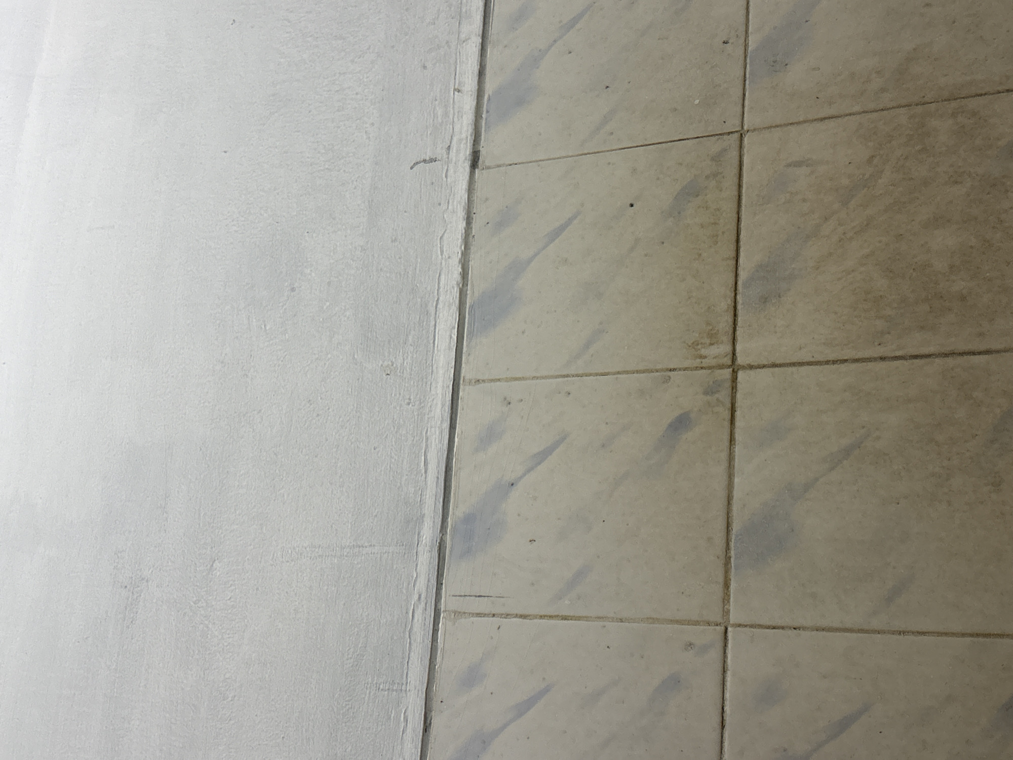 A close-up, eye-level shot shows a tiled floor meeting a white, textured wall. The floor is composed of square, light-brown tiles with visible grout lines. The tiles appear somewhat dirty, with dark, elongated smudges that suggest shadows or water stains. The wall above the floor is painted white, exhibiting a rough, uneven texture with subtle variations in shading. A thin, dark line runs horizontally along the top edge of the tiled area, separating it from the wall. There are no people, animals, or other discernible objects present in the scene. The lighting is even, casting no harsh shadows. The image captures a mundane architectural detail, possibly a corner or floor-to-wall transition, in an urban setting.