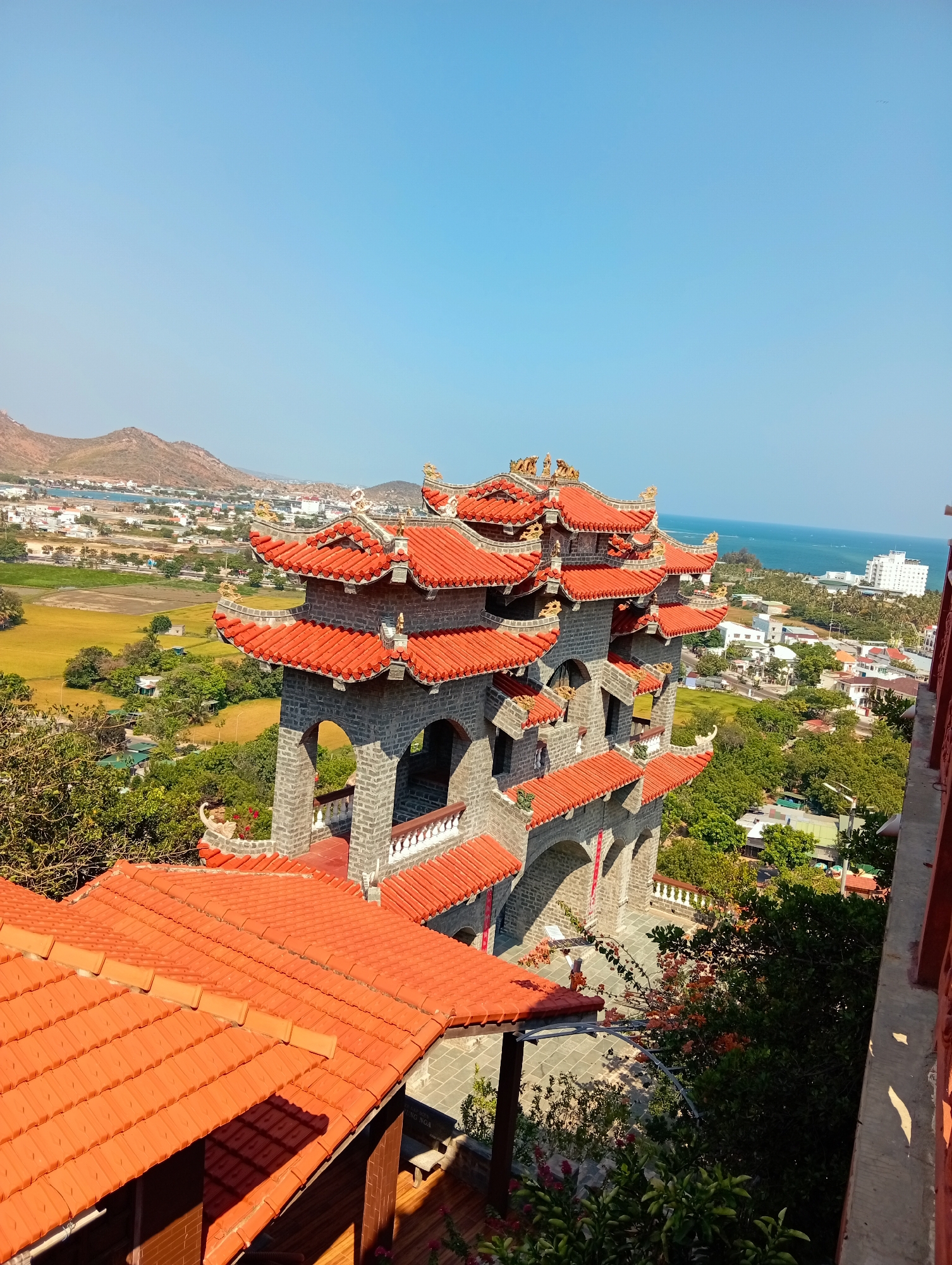This high-angle photograph captures a breathtaking panoramic view of a multi-tiered, pagoda-like structure and the surrounding coastal landscape in Khánh Hải, Vietnam, on a clear, sunny day. The dominant subject is a grand building featuring a gray, stone-like facade and distinctive bright orange-red tiled roofs. Each of its multiple levels boasts curved, ornate eaves, adorned with intricate golden decorations, possibly depicting mythical figures, along the ridges and corners. The structure includes arched openings and balconies with white railings.
The vantage point reveals a diverse natural and urban setting. To the left, a range of arid, brownish mountains stretches into the distance. Below them, expansive yellow-green agricultural fields, likely rice paddies, are visible, dotted with patches of lush green trees and scattered small buildings. A bustling town or city occupies the middle ground, with numerous buildings extending towards the sea. To the right, the calm, deep blue ocean meets the horizon, with several white buildings lining the distant coastline.
The image conveys a sense of tranquility and cultural splendor. No people are visible, allowing the viewer to focus on the architectural details and the vast landscape. The bright sunlight and clear blue sky suggest it is midday or early afternoon, with strong, defined shadows. Faint, unreadable text in what appears to be Vietnamese is barely discernible on a sign in the lower right corner.