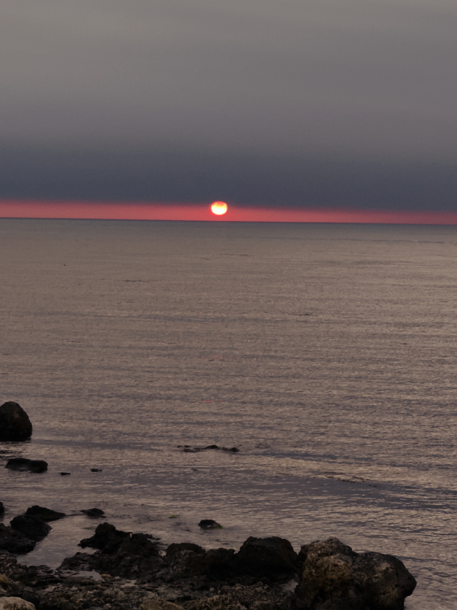The image shows a sunset over the Black Sea. The sun appears as a red orb, setting on the horizon. The sky is a gradient, darker grey above, transitioning to red near the horizon where the sun is setting. The sea is calm with gentle waves. Rocks and pebbles are visible in the foreground, along the shoreline. The location is likely the city of Sevastopol, Ukraine. No people are visible in the scene.