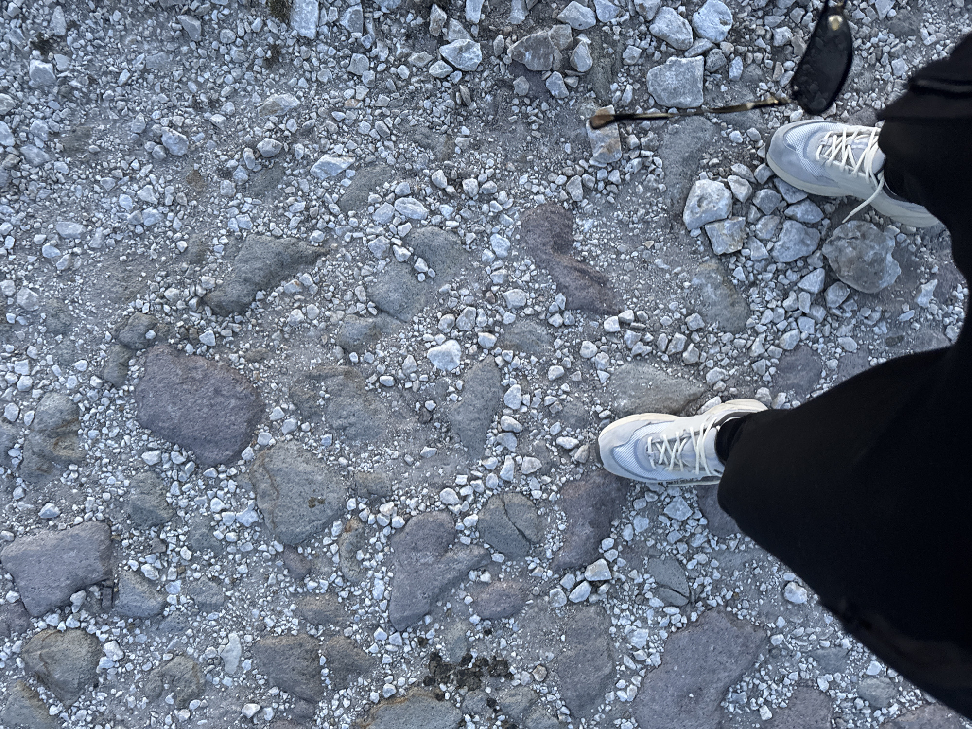 The image shows a first-person perspective of someone standing on a rocky, uneven surface. Their feet, clad in white sneakers and black pants, are visible in the foreground. To the right of the right foot, a pair of dark sunglasses and a thin, dark object, possibly a walking stick or cane, are resting on the ground. The ground is composed of a mix of small grey pebbles and larger, irregularly shaped grey and purplish stones. There are no other people or structures visible in the frame. The environment appears to be outdoors, with the lighting suggesting it is daytime. The location is identified as the city of Gela, Bulgaria, although there are no specific landmarks or indicators in the image to confirm this.