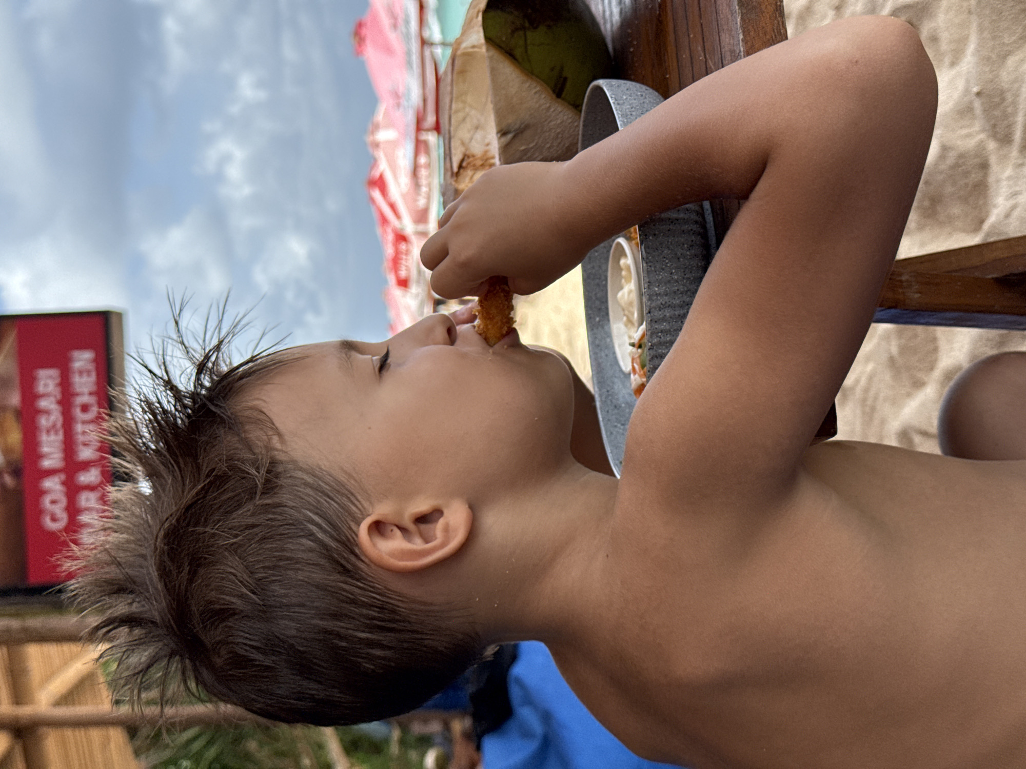 A young, shirtless child is eating a fried food item, possibly a shrimp, at an outdoor table in Kuta Selatan, Indonesia. A coconut drink sits on the table. The child is positioned in profile, facing left, with their mouth open to take a bite. A gray bowl with a dip rests near the child's hand. In the background, a sign reads "GOA MESARI BAR & KITCHEN." Beyond the table, a beach scene is visible with sand and partially obscured umbrellas and structures. The sky is overcast.