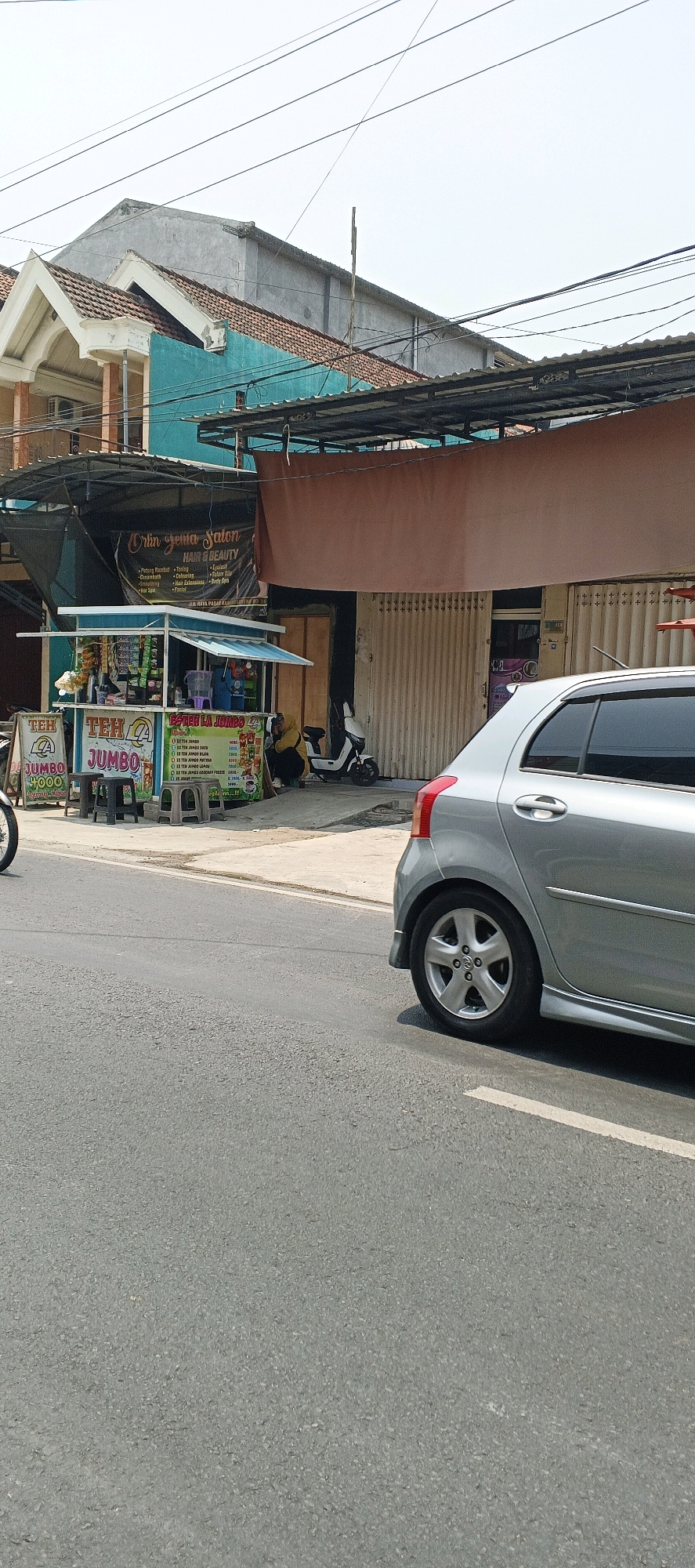 In the city of Sumberwudi, Indonesia, this image captures a street scene featuring a small food stall, a silver car, a scooter, and buildings under a bright sky. The food stall, adorned with colorful signage advertising "TEH JUMBO," is positioned on the left side of the frame. It displays various drinks and snacks, with several plastic stools and a bicycle wheel visible nearby. A person wearing a yellow hijab is seated next to the stall, interacting with the vendor. To the right of the stall, a white scooter is parked.
Behind the stall, a building with a teal facade and a tiled roof is visible, partially obscured by a brown tarp that extends over a storefront with closed metal shutters. A sign for "Orlin Jemia Salon HAIR & BEAUTY" is displayed on the upper part of this building. Further back, a more substantial building with a grey concrete exterior and a pitched roof is seen.
In the foreground, a silver car is parked on the right side of the road, with its rear visible. The road surface is asphalt, marked with a white line on the right side. Overhead, power lines crisscross the sky, indicating an urban or semi-urban environment. The overall scene suggests a typical day in a commercial area.