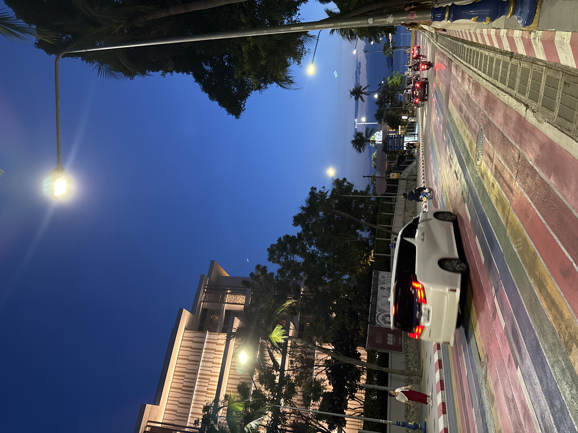 The image depicts a street scene in Pattaya City, Thailand, at dusk. A white minivan is driving across a pedestrian crossing painted with rainbow stripes. The street is lined with palm trees and modern buildings. Streetlights illuminate the scene, and other vehicles, including motorbikes, are visible in the background. A woman in a red dress and white cardigan is standing on the sidewalk. The sky is a deep blue, transitioning from twilight.