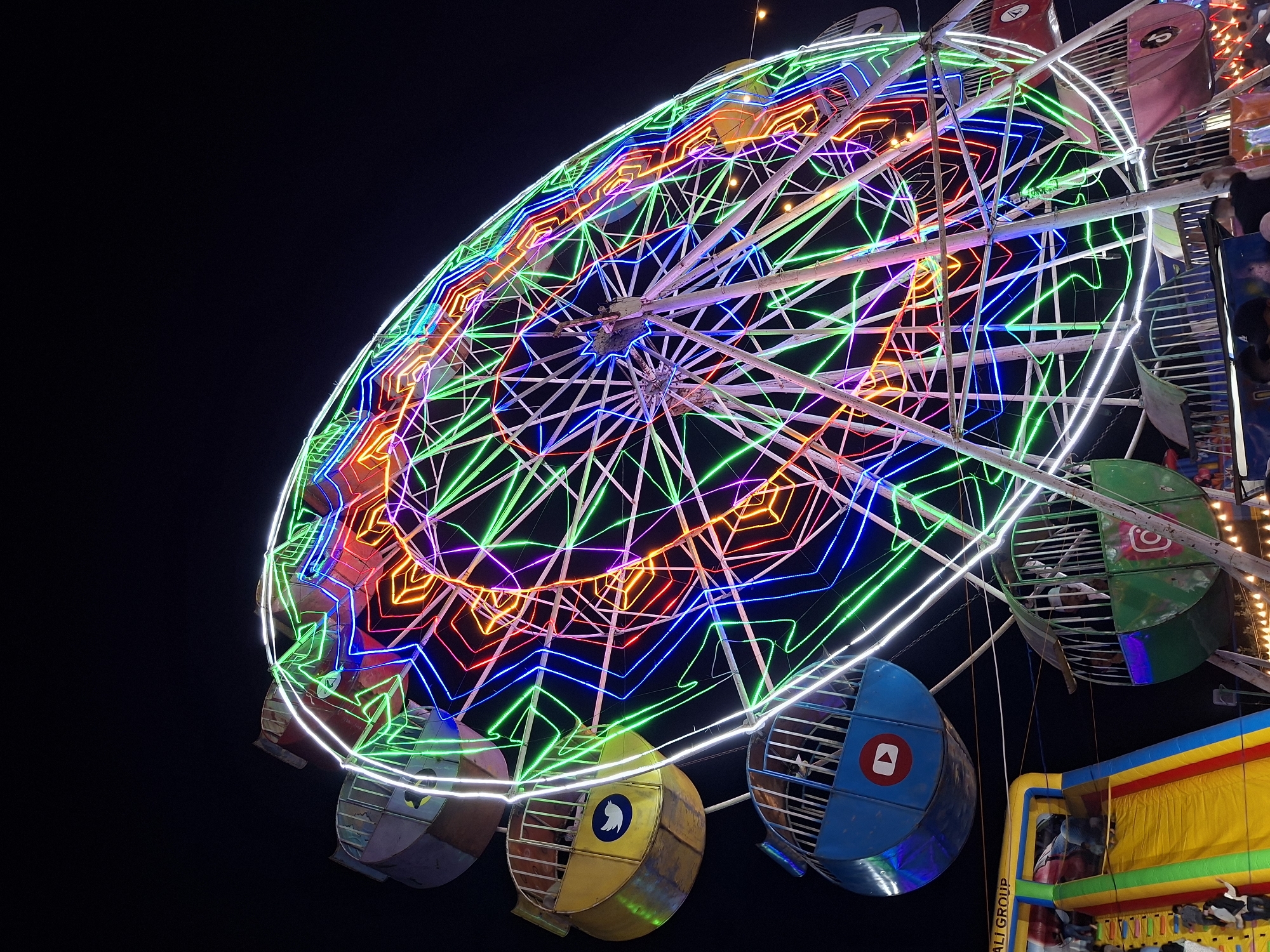 The image depicts a brightly lit Ferris wheel against a dark night sky. The Ferris wheel is adorned with multiple colored neon lights – red, orange, yellow, green, blue, and white. The spokes of the wheel are illuminated with white lights. The passenger cars are painted in different colors including yellow, blue, and green, each bearing a distinctive circular logo.
The image was taken in Geulumpangsulu Timu, Indonesia. The context indicates that the Ferris wheel is part of a carnival or fairground, as indicated by the bright lights and multiple colors. The image captures the wheel at night. The lower right corner of the image shows parts of the ride and some people are in the cars.