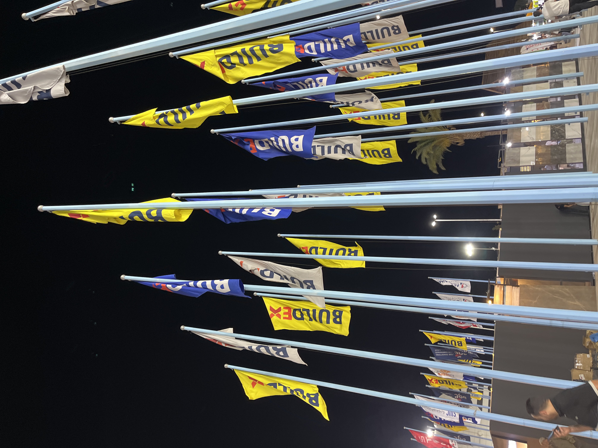 The image shows an outdoor event at night in Al-Ghizlaniya, Syria. A multitude of flags are displayed on tall poles. Many of the flags are yellow and blue, with text that appears to be "BUILDEX" and "BUILD". Some white flags with blue lettering are also present. In the background, there are buildings and lighting fixtures. Several people are visible in the foreground, some walking and one standing while looking at their phone. The environment appears to be a public space or exhibition area, with a palm tree visible in the mid-ground.