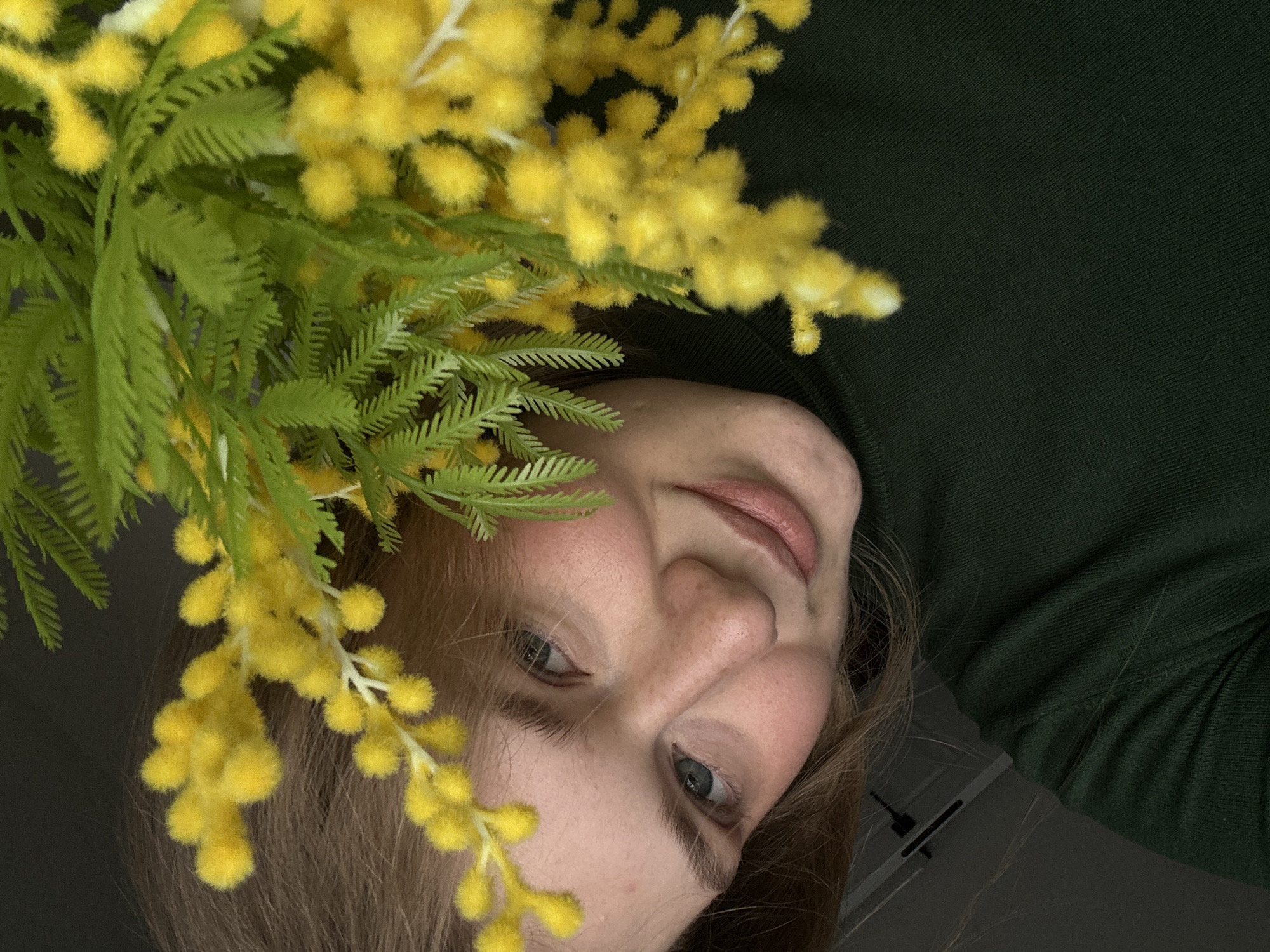 The image is a close-up, slightly tilted view of a woman with fair skin, light eyes, and light brown hair, alongside a bouquet of yellow mimosa flowers. The woman is wearing a dark green turtleneck sweater. The mimosa flowers are positioned on the left side of the image, partially obscuring the woman's face. The background is a dark, indistinct space. It is not possible to confirm the location of the image as Kazan, Russia with the information provided.