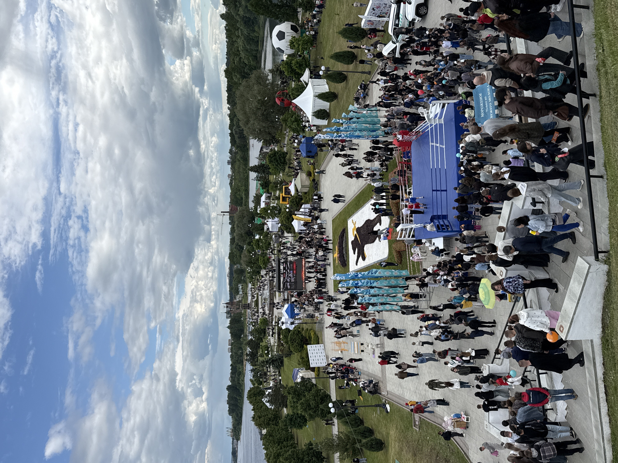 The image shows an aerial view of an event in the city of Шебунино, Russia. Numerous people are gathered in a park-like setting near a body of water. Several tents and booths are set up, including what appears to be a boxing ring. A large crowd is assembled to observe the event. In the background, an enormous soccer ball is visible. The sky is partly cloudy.