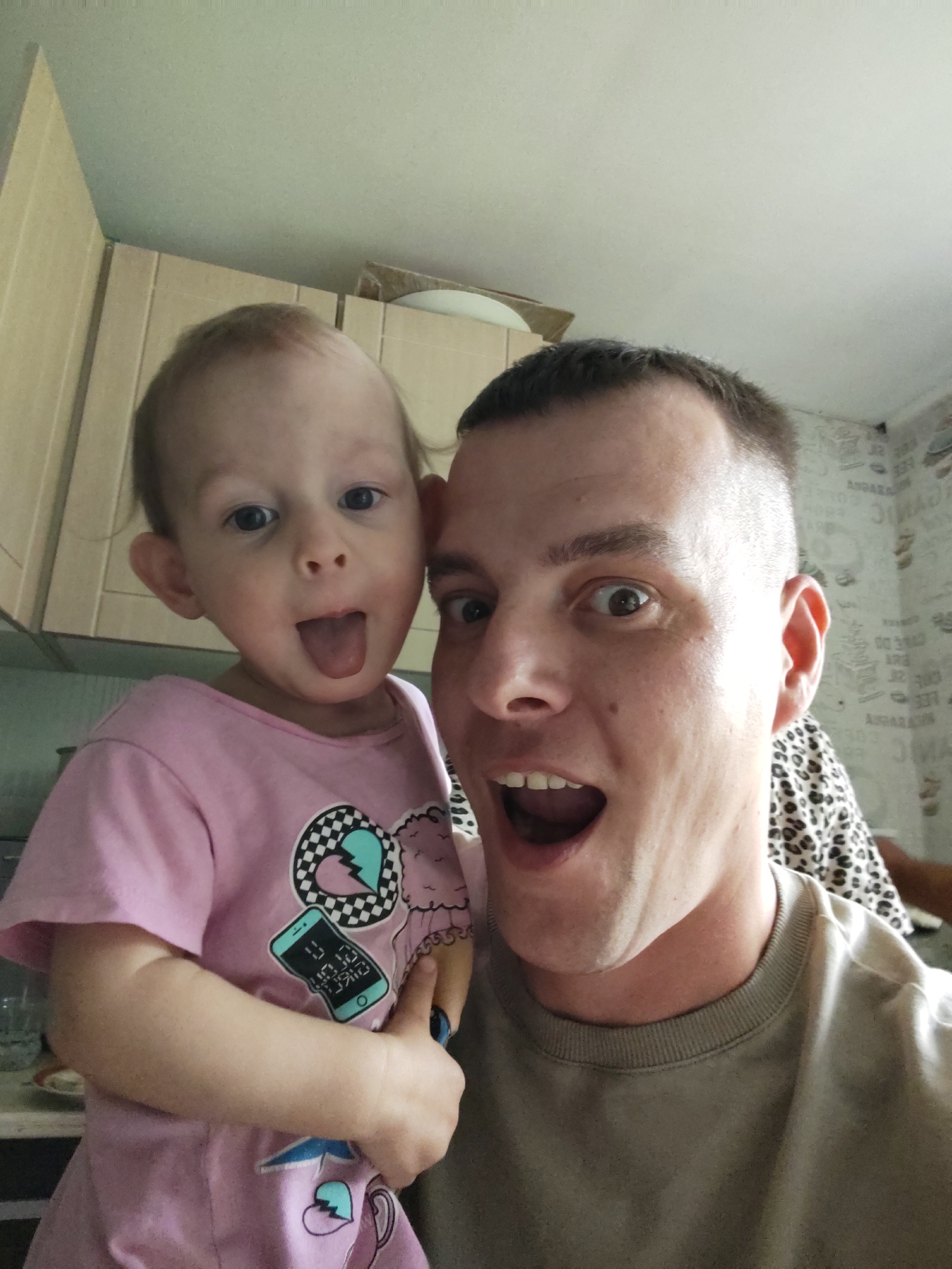 This close-up selfie captures an adult male and a young child, likely a girl, engaging in a playful moment indoors in Nizhnyaya Gar', Russia. Both subjects are looking directly at the camera with exaggerated, joyful expressions, suggesting a lighthearted interaction during the daytime.
The child, positioned on the left side of the frame, has wide eyes and is sticking out her tongue in a mischievous gesture. She is wearing a pink short-sleeved t-shirt decorated with several graphic designs, including a black and white checkered heart, a broken blue heart, and a stylized teal smartphone icon featuring illegible text. Her right hand, holding a small blue object, is visible near her hip.
The adult male, on the right, also has wide, surprised-looking eyes and an open mouth. He has a short, close-cropped haircut. He is wearing a plain, olive-green crew-neck sweatshirt.
The background indicates a home interior, possibly a kitchen or utility area. Light-colored wooden cabinets or cupboards are visible behind the child and in the upper left corner. On the right, behind the man, there is wallpaper with a repetitive pattern of indistinct, blurry dark text-like elements on a light background, including shapes resembling letters or numbers such as "AZAZA." The overall mood of the image is cheerful and spontaneous.