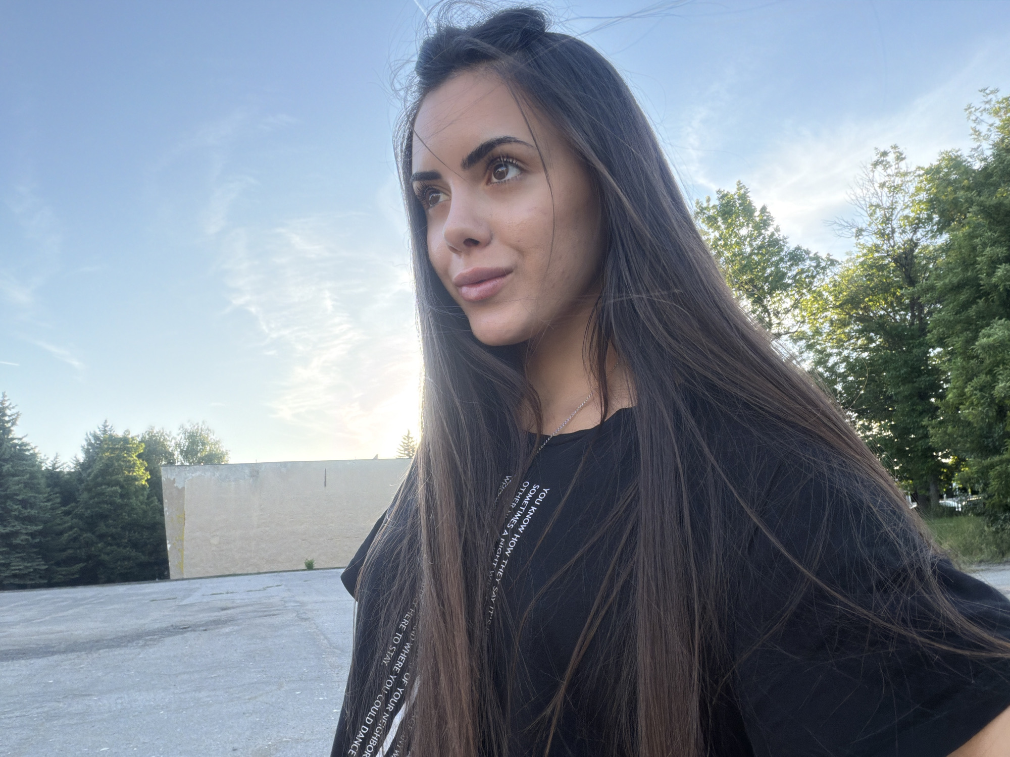 The image shows a young woman with long, dark brown hair, wearing a black t-shirt with white text on the sleeve and front. She is looking off to the side, with a slight smile. The background includes a clear blue sky with some light clouds, trees, and a tan building. The ground is asphalt. The text on her shirt mentions "dance" and "neighbor".
While the image contains elements that are also present in the city of Sofia, Bulgaria, there is nothing in this image to specifically indicate the location as Sofia, Bulgaria.