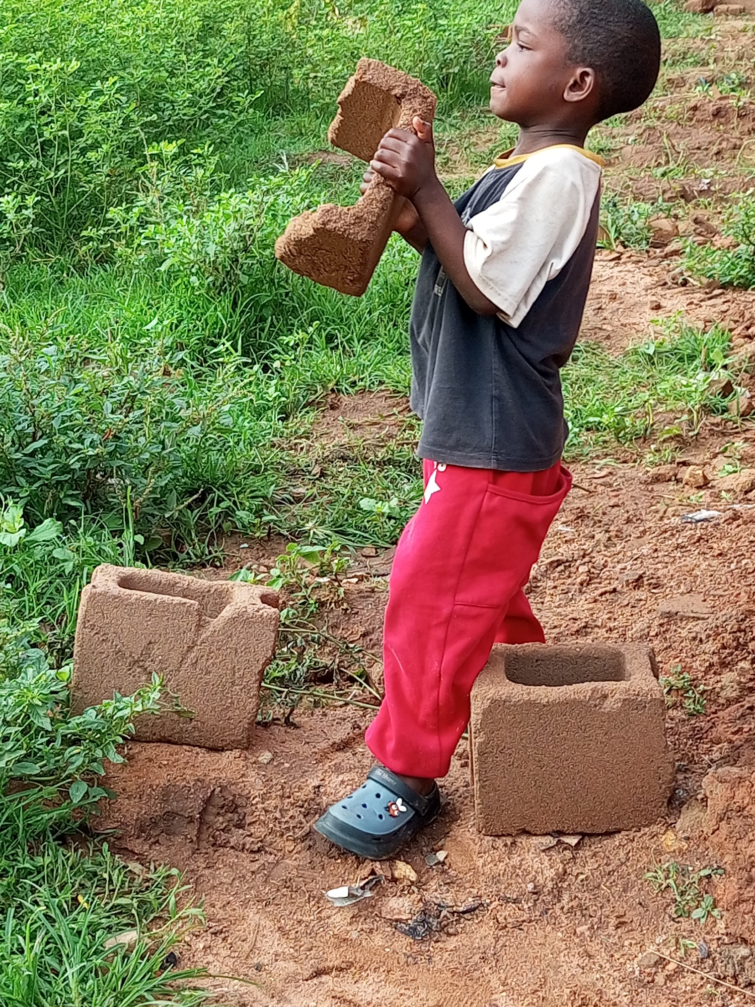 In Tempure, Nigeria, a young boy stands on a dirt path surrounded by greenery. He is wearing a black and white t-shirt and red pants. In his hands, he holds a brown object that appears to be made of clay or mud. Beside him on the ground are two identical brown blocks, also made of clay or mud, and a single blue shoe. The boy is looking upwards and to the right. The scene suggests a rural or developing area.
