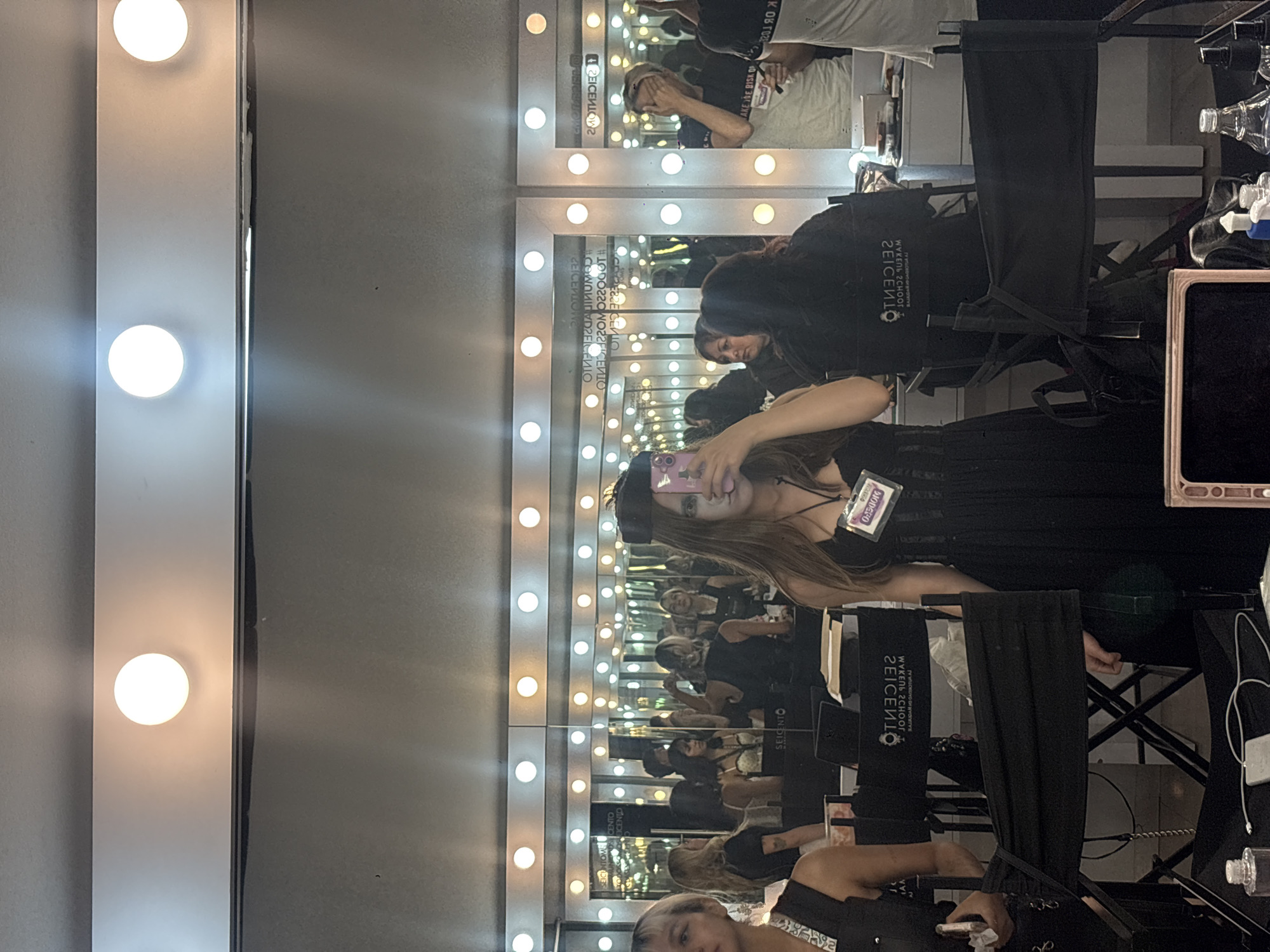 The image shows a group of individuals in a brightly lit room, likely a makeup studio or backstage area, possibly at the SEICENTO MAKEUP SCHOOL in Mexico City, Mexico, indicated by text on some of the chairs. Several lighted mirrors, each with multiple illuminated bulbs, reflect the scene and the people within it.
A woman in the foreground is holding a phone and taking a picture of herself in one of the mirrors. Other people in the scene appear to be makeup artists or students. They are seated in director-style chairs and attending to clients.
The environment is busy and focused, suggesting a preparation session for a performance or photoshoot. The scene is cluttered with makeup equipment, bottles, and personal items on a table.