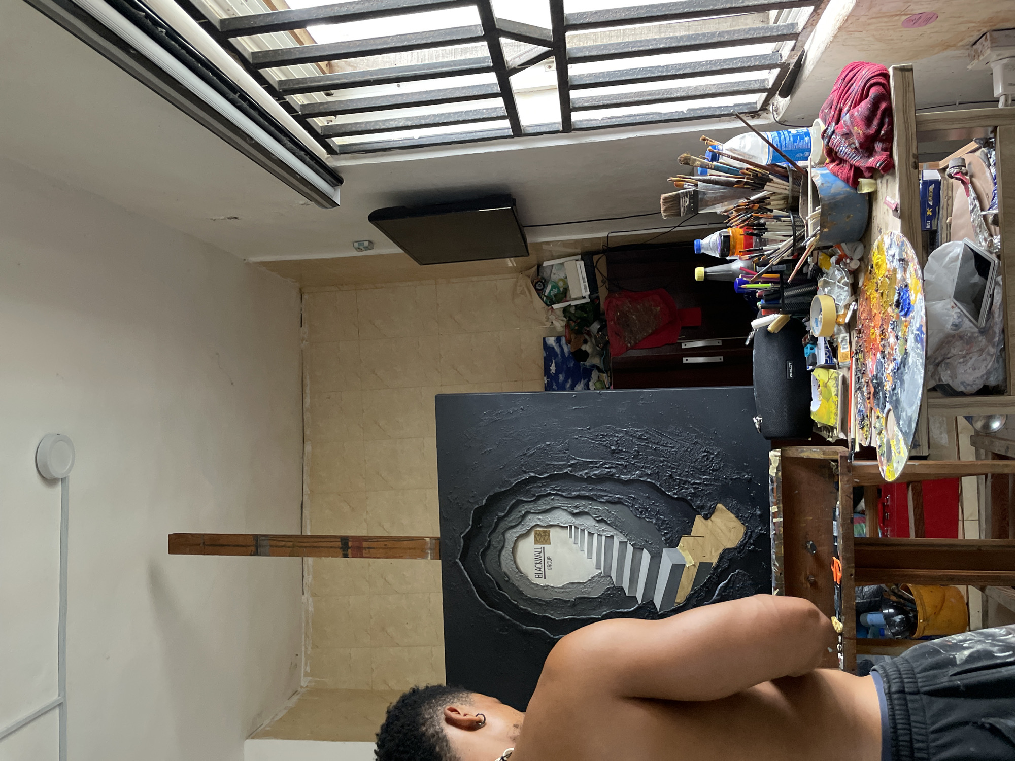 An undated, medium shot, eye-level, indoor, natural-light photograph captures an artist, a shirtless Black man with short curly hair and an earring, standing in an artist's studio in Ado, Nigeria. The artist is positioned to the left of the frame, facing away from the viewer and towards a large, textured painting on an easel. The painting, predominantly black, depicts a tunnel-like entrance with a set of grey and gold stairs leading upwards into a brightly lit opening that displays the text "BLACKWALL GROUP."
The studio appears somewhat cluttered, with art supplies scattered across a wooden easel and a table. Visible on the easel are brushes, tubes of paint, and what appears to be a palette knife. A palette laden with colorful paints is on the table, alongside various bottles, containers, and a black speaker. A red and white plaid cloth is draped over the table. In the background, a window with metal bars is visible on the right, letting in natural light. A small black television is mounted on the wall to the left of the window. The walls are tiled with a light-colored mosaic pattern, and the ceiling is white with some visible signs of wear. The overall impression is of a working artist's space, filled with the tools and materials of creation.