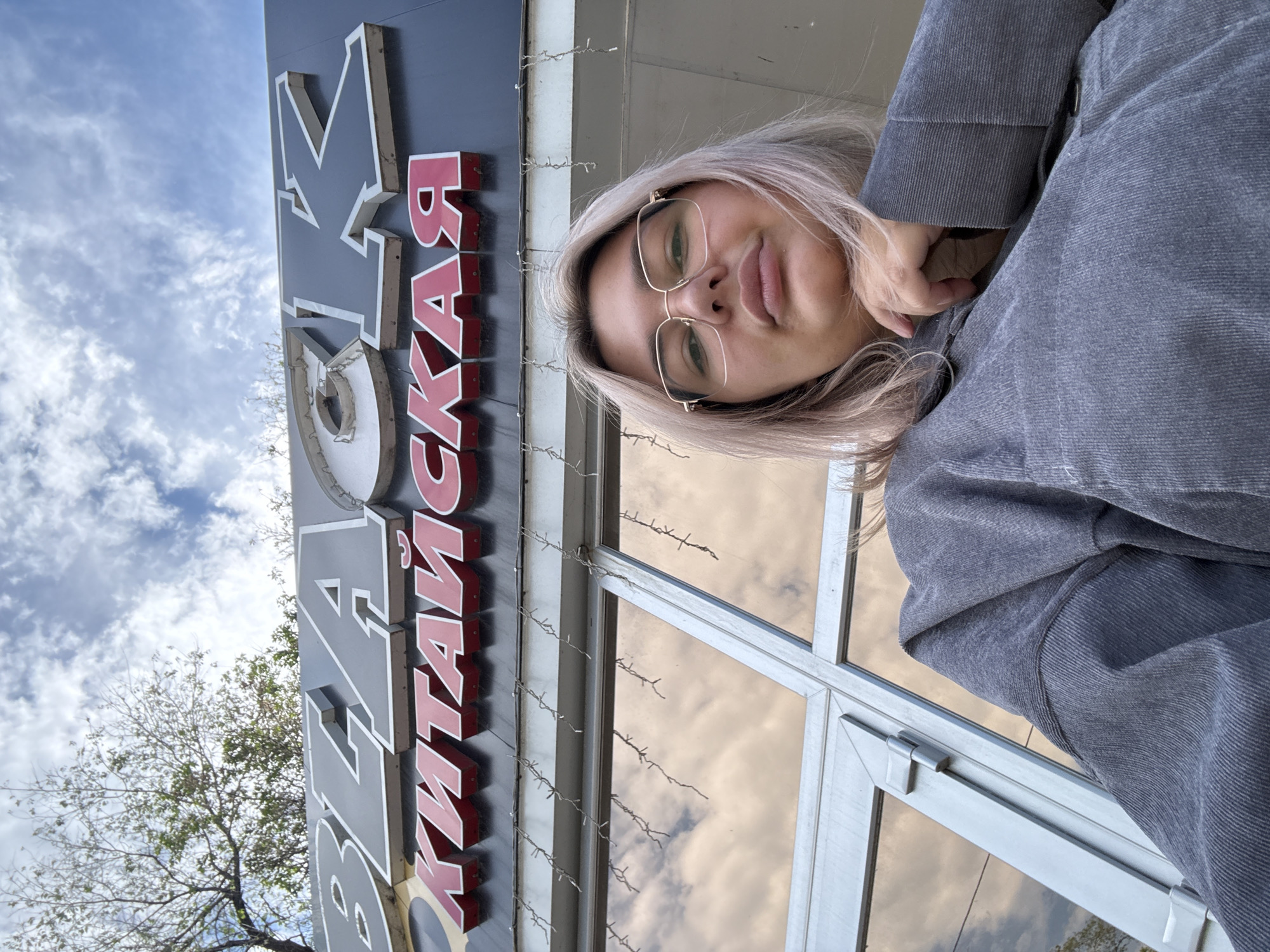 A low-angle, outdoor, wide shot shows a young woman with light blonde hair and glasses taking a selfie in front of a building. The building's facade features a large, dark sign with white lettering that spells "BLACK" across the top, and a red sign below with Cyrillic lettering. The woman, positioned in the lower right of the frame, wears a gray hoodie. Her left hand is resting on her chin. Behind her, the building has large, reflective windows and is adorned with small, string lights. The sky above is partly cloudy. The sign's lettering and the word "КИТАЙСКАЯ" (which translates to "CHINESE") suggest the establishment is a Chinese restaurant. Birobidzhan, Russia, is known for its significant Jewish population and its history as an autonomous oblast within the Soviet Union.